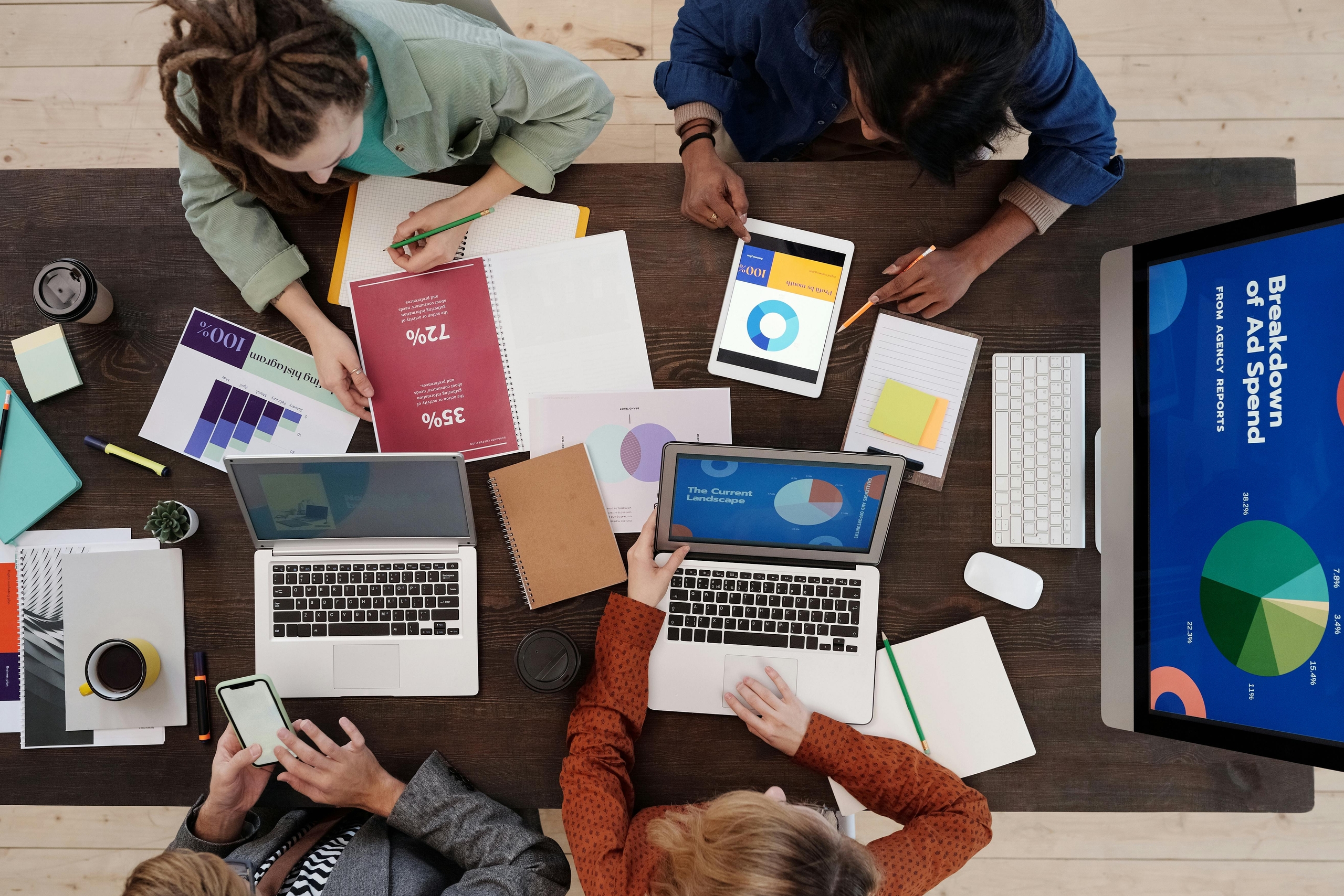 A diverse group collaborating over laptops and tablets on a wooden table, with various documents and notes spread out around them. The screen displays colorful data visualizations, highlighting teamwork in community service and civic leadership projects.