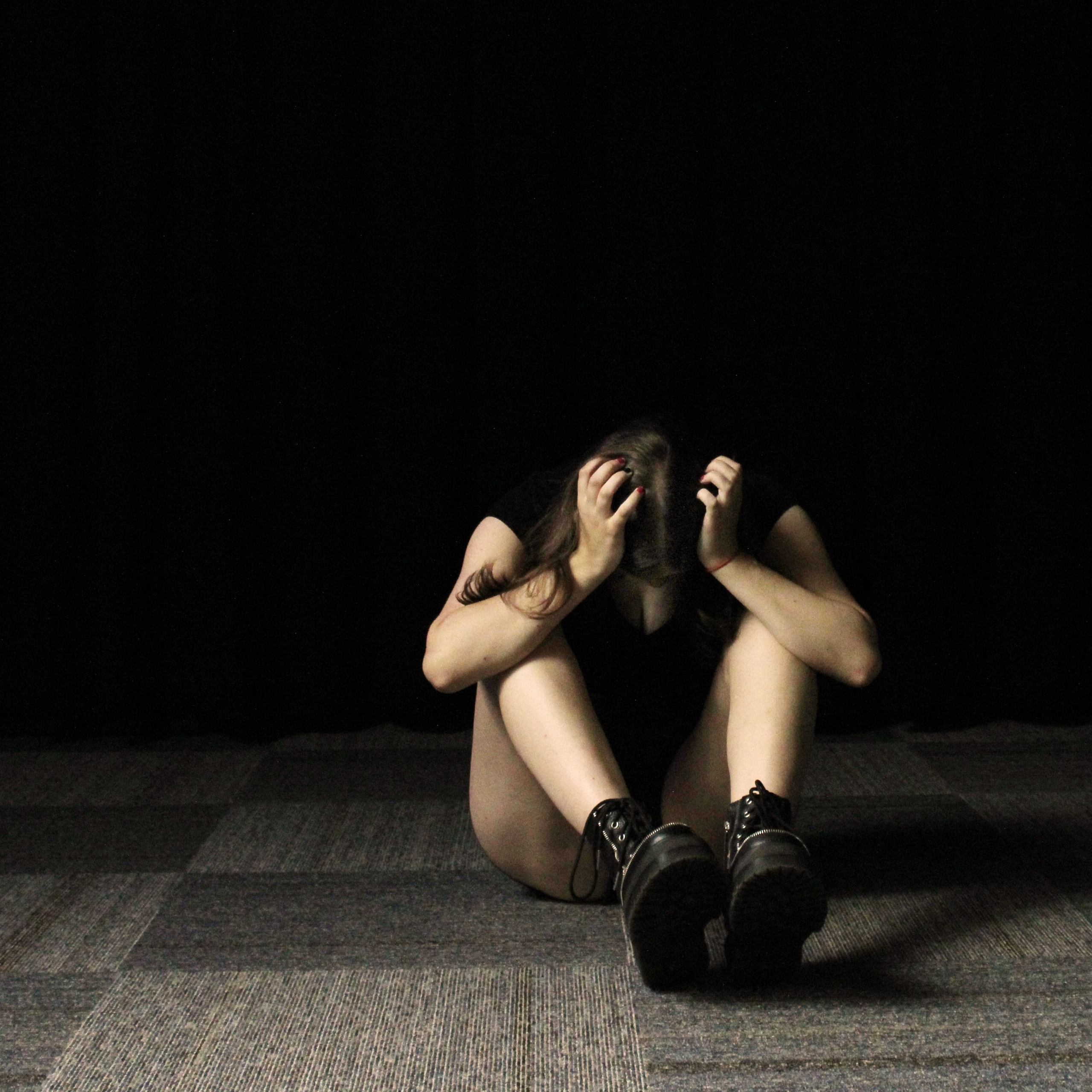 A person sits on the floor in a dimly lit space, with their head in their hands, conveying a sense of distress or contemplation. The setting features textured gray carpet and a black background, emphasizing the individual's isolation and emotions.