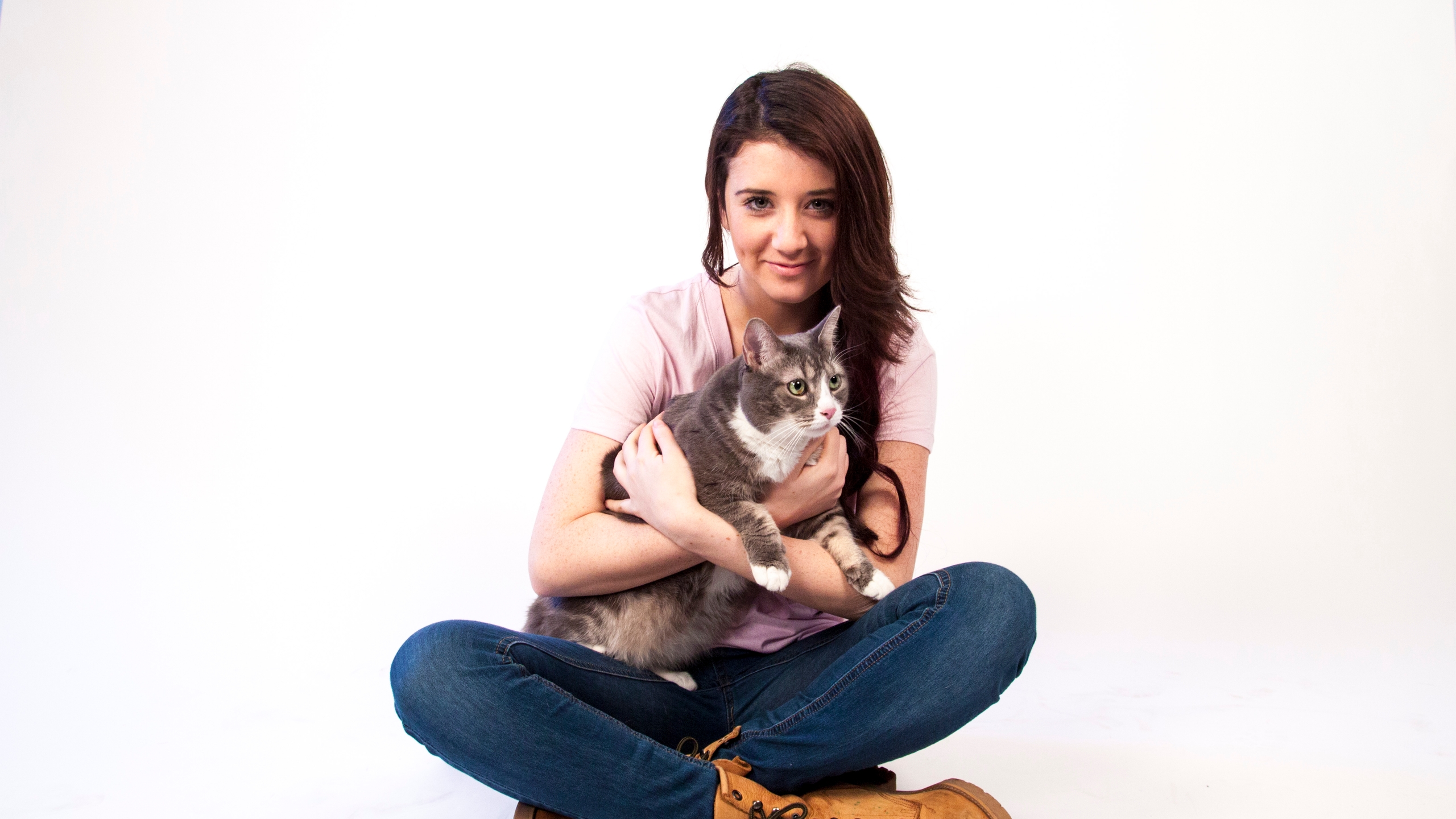 A young person with long hair, wearing jeans and a pink shirt, seated cross-legged while hugging a cute tabby cat