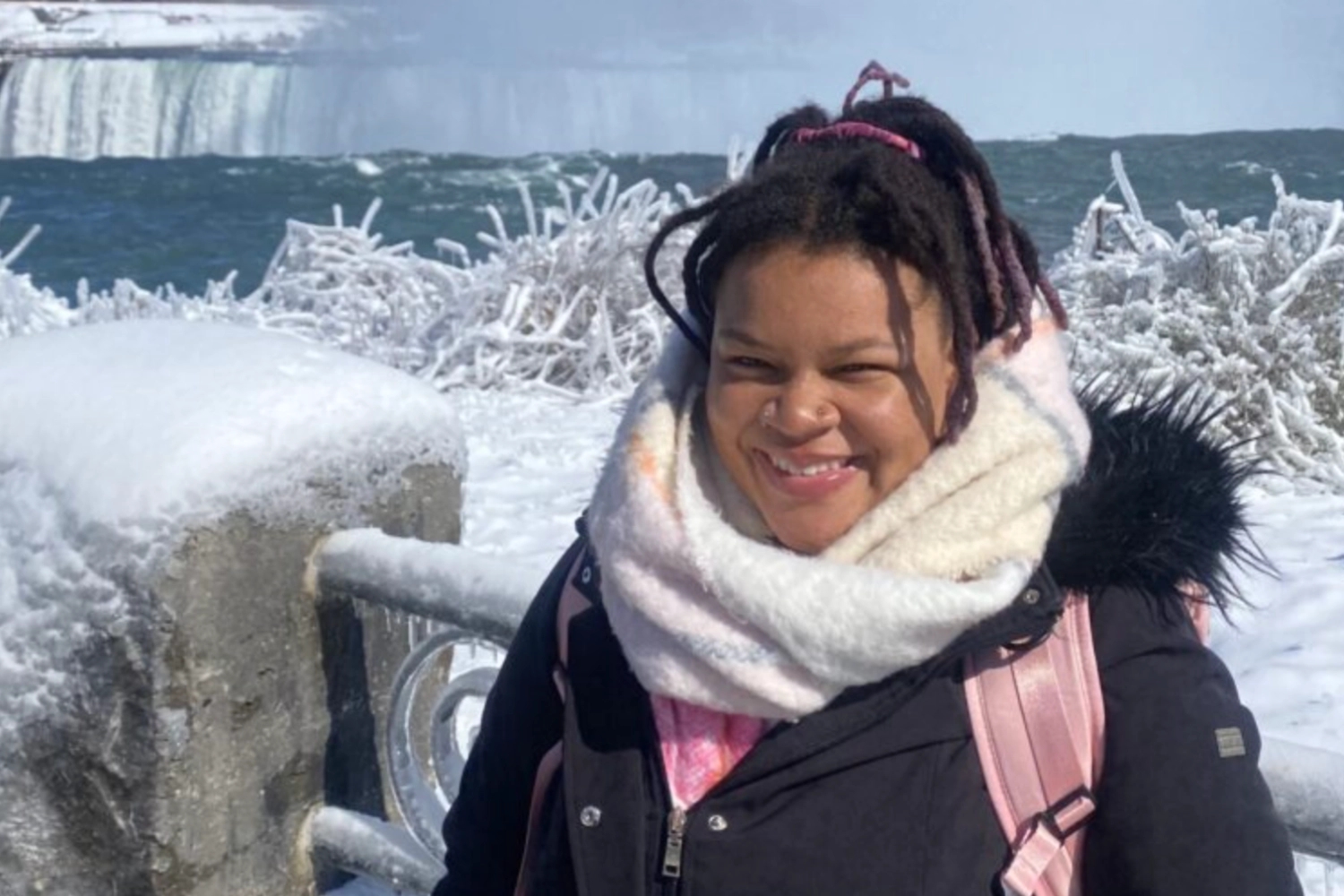 A smiling individual stands near a snowy railing with Niagara Falls in the background, surrounded by icy scenery. They are dressed warmly in a black jacket and scarf, ready to engage in community service and civic learning opportunities.