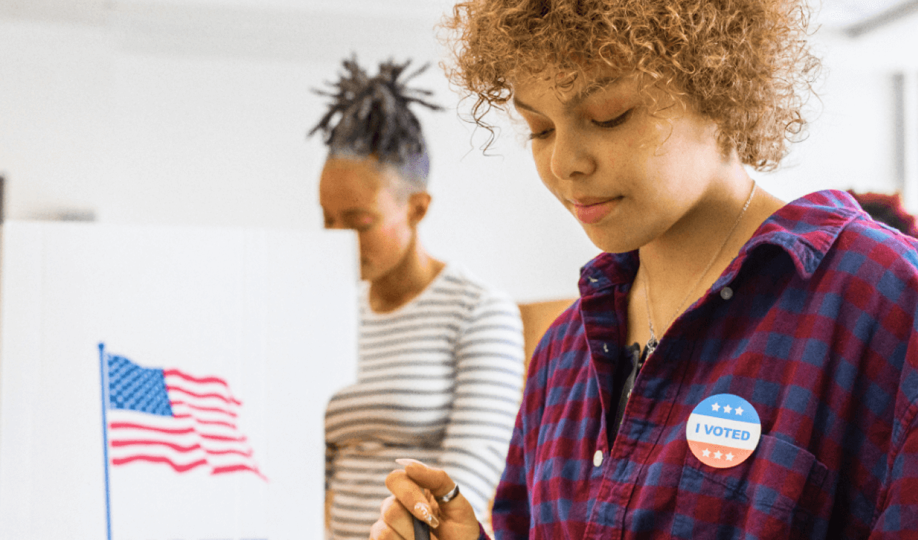 A young person casting their ballot, wearing an 'I Voted' sticker; a neighbor casts their ballot in the background, in a voting booth adorned with an American flag sticker 