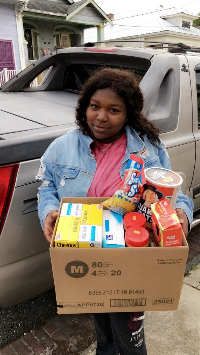 An individual stands next to a vehicle, holding a cardboard box filled with various food items including cereals, snacks, and sauces. This scene illustrates community service efforts through volunteer activities aimed at addressing local food needs.