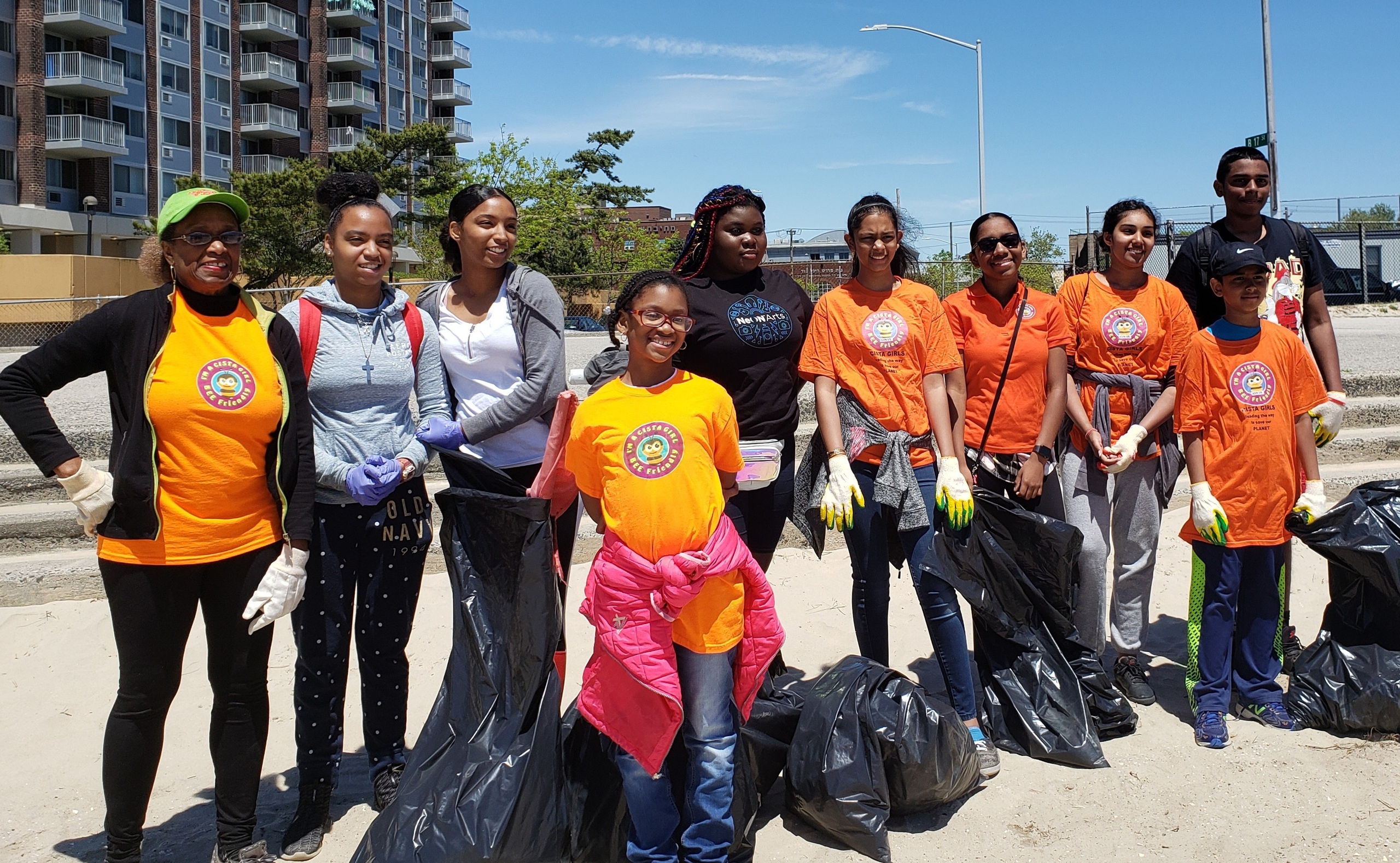A diverse group of volunteers, wearing orange t-shirts and gloves, smiles while holding trash bags during a community clean-up event. The background features a sunny day and an urban setting, emphasizing engagement in volunteer and civic initiatives.
