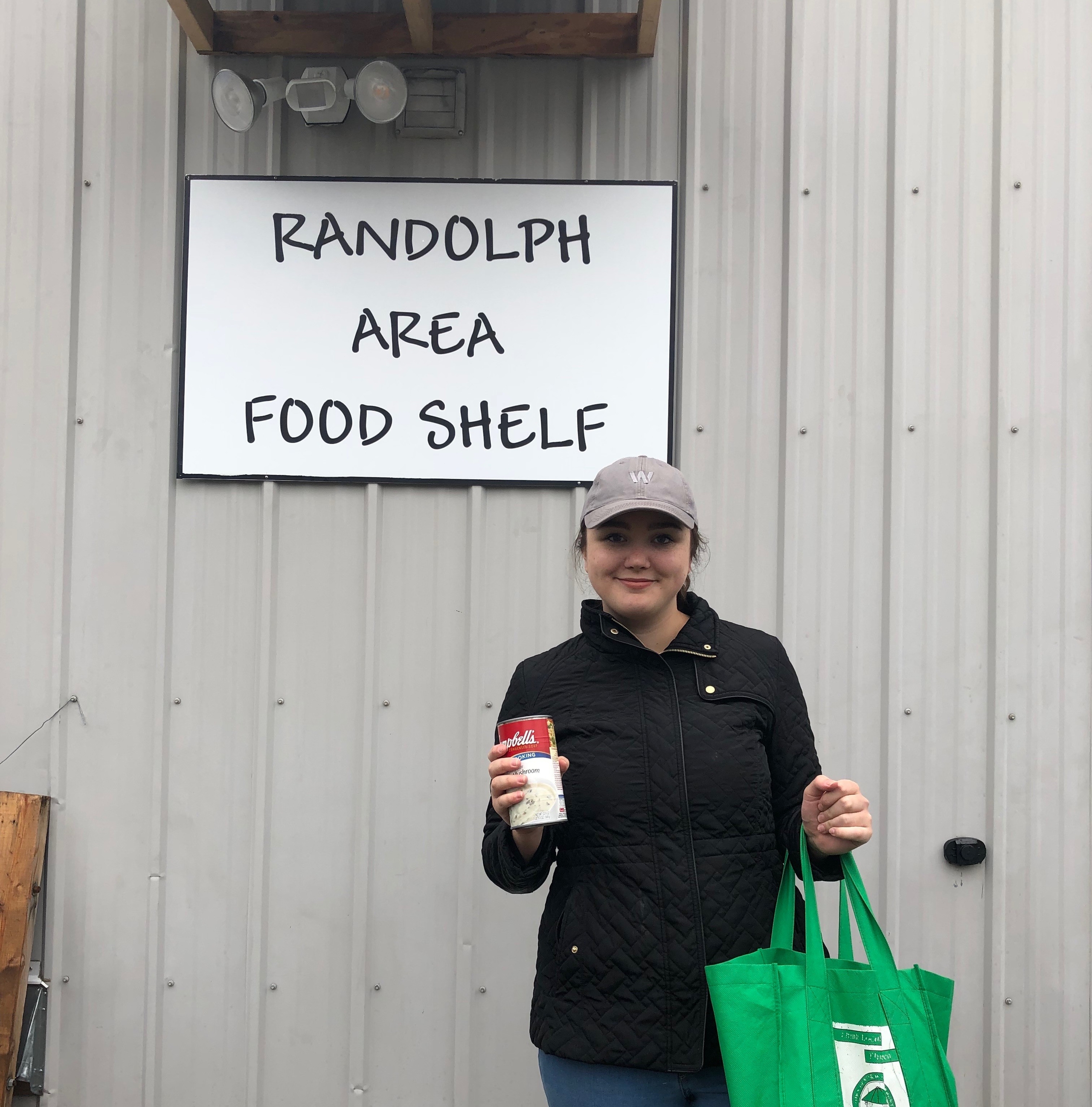 A person stands in front of the Randolph Area Food Shelf holding a can of soup in one hand and a green reusable bag in the other. The sign above displays the name of the food shelf, emphasizing community engagement and support.