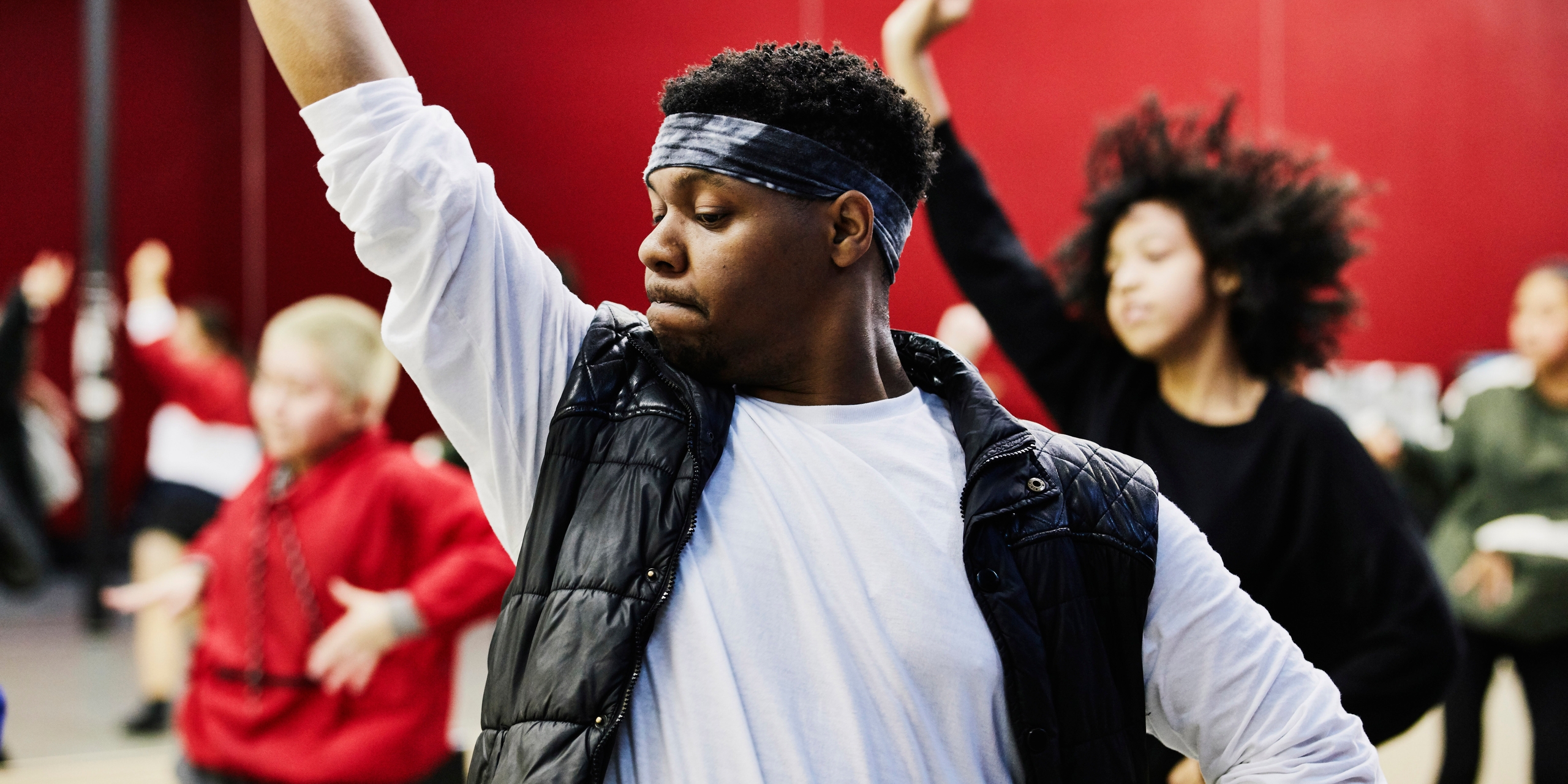 A diverse group of individuals participates in a dance class, focusing on movement and rhythm. One person in the foreground raises their arm confidently, showcasing the joy of community engagement and creativity.