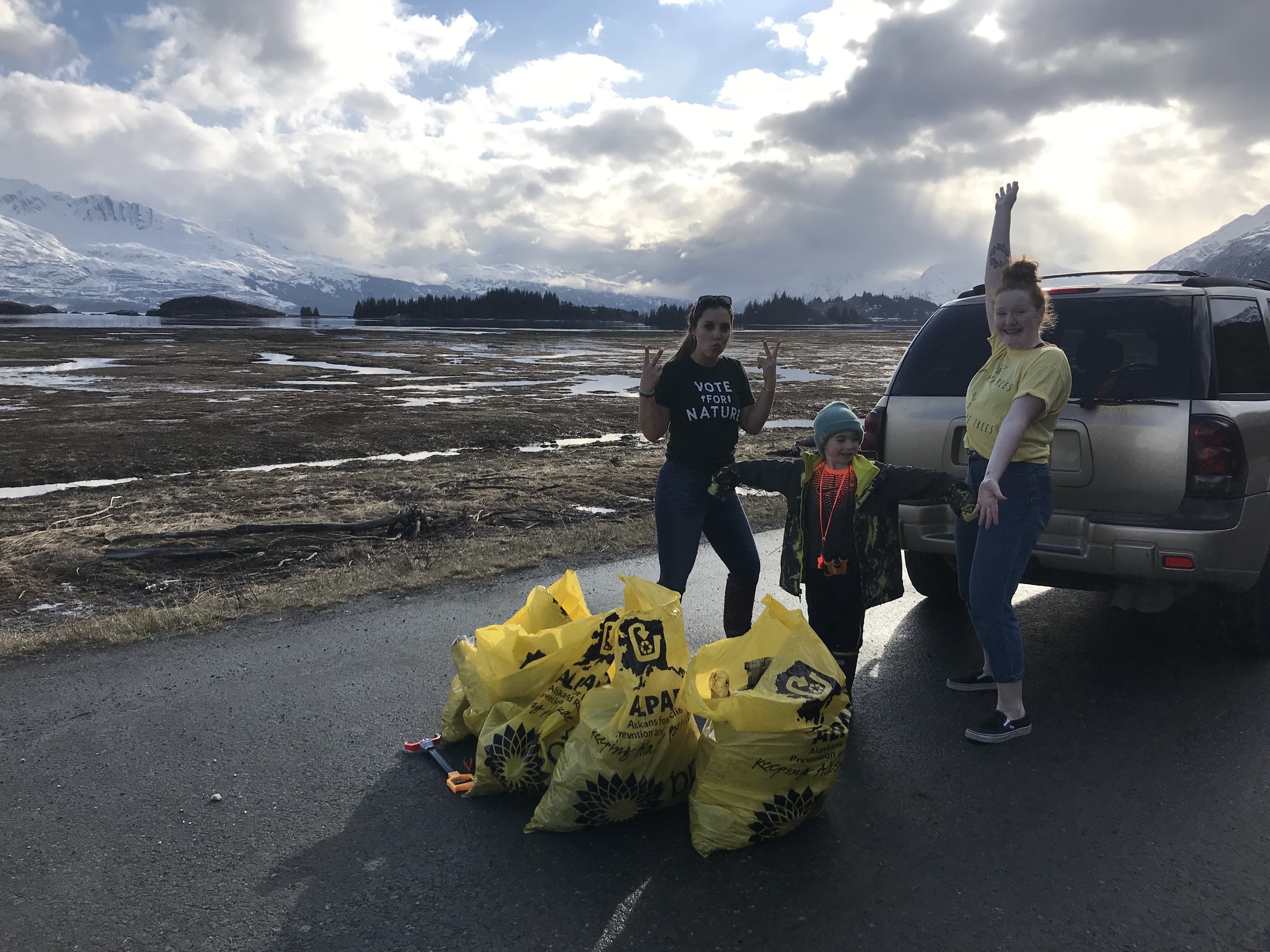 A group of three volunteers stand beside a pile of yellow trash bags filled with collected litter, set against a backdrop of mountains and cloudy skies. The individuals display cheerful expressions while participating in a community service project to promote environmental awareness.