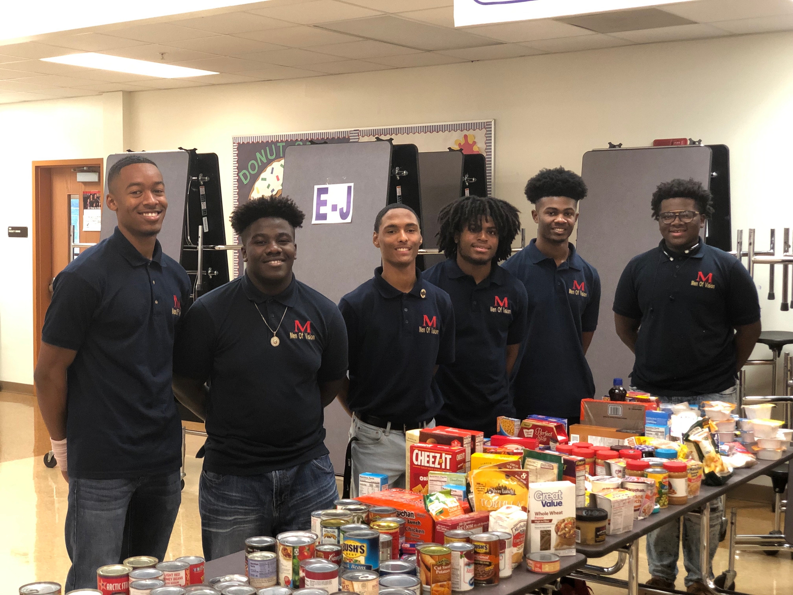 A group of six community volunteers stands proudly behind a table filled with various food items for a donation drive. All volunteers wear navy blue polo shirts with the logo 'Men of Valor', showcasing their commitment to community service and civic engagement.