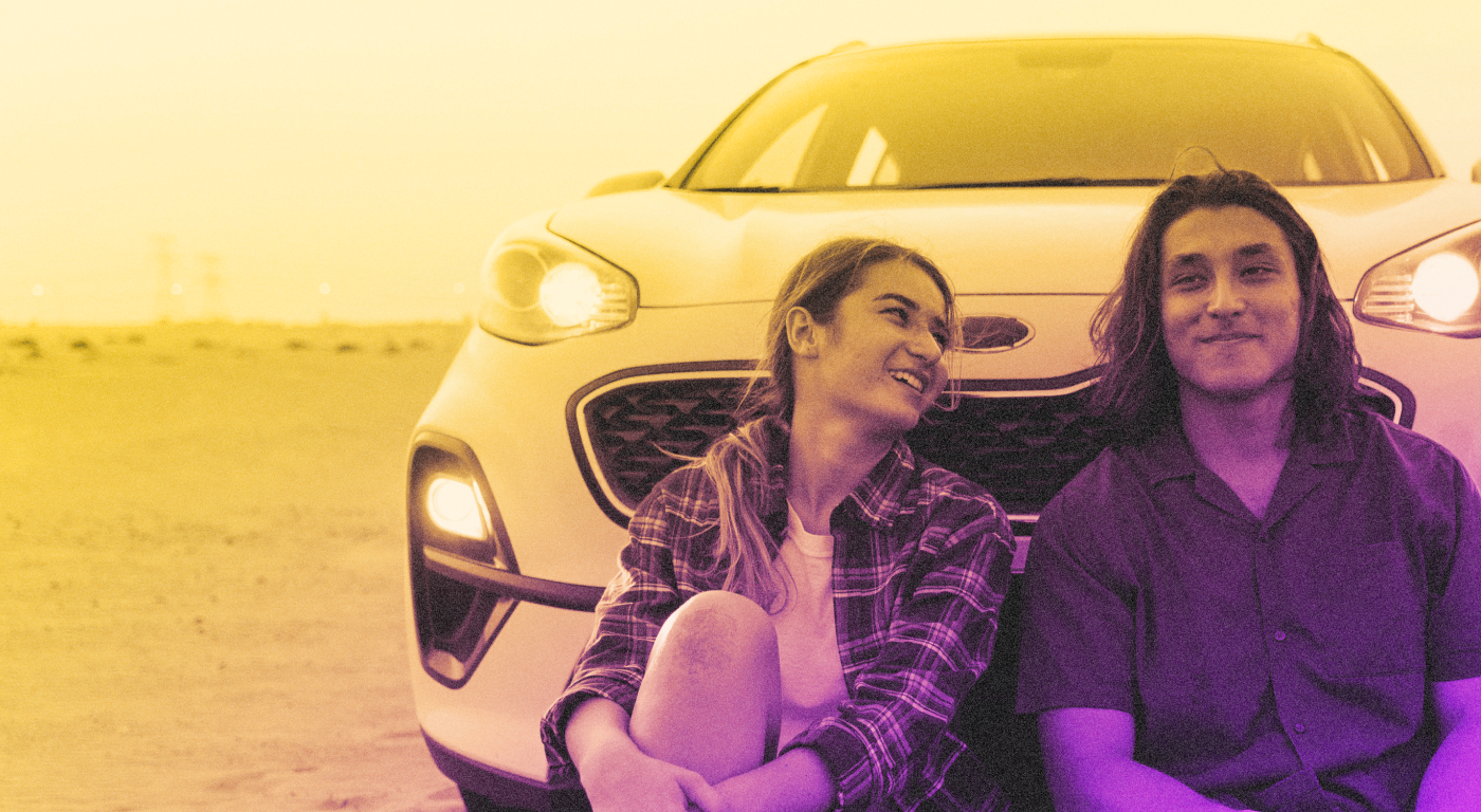 A pair of young people seated on the ground, leaning agains the front grill of a car, talking and laughing