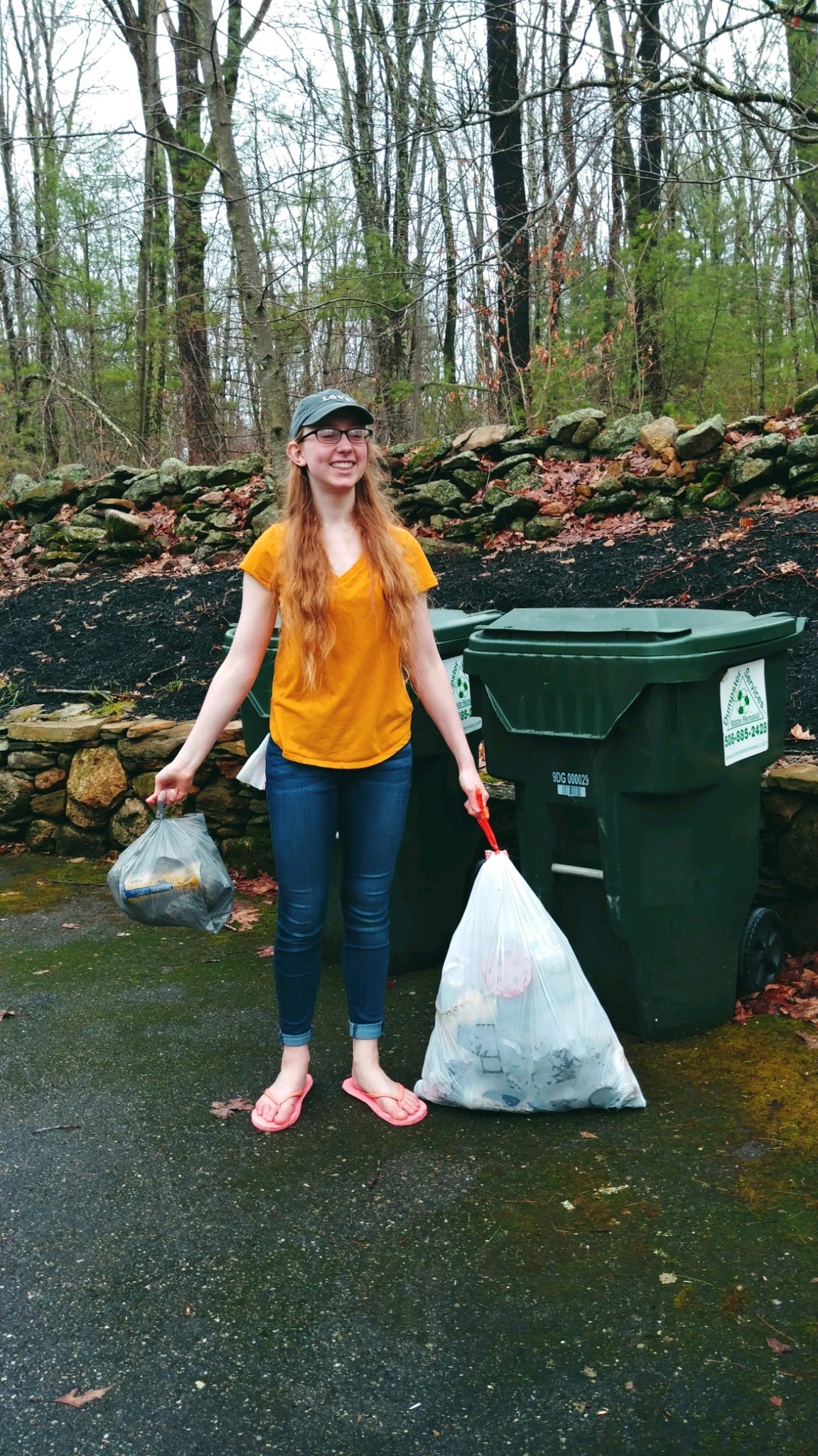 A smiling individual stands outdoors, holding two gray bags filled with collected trash, ready for disposal next to green recycling bins. This scene highlights community service and environmental volunteering efforts, promoting civic engagement.