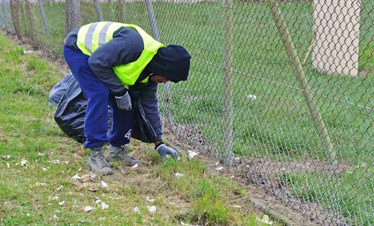 A person wearing a bright green safety vest and gloves is picking up litter along a fence in a grassy area, promoting community cleanup efforts. This image highlights volunteer opportunities focused on environmental stewardship and civic engagement.