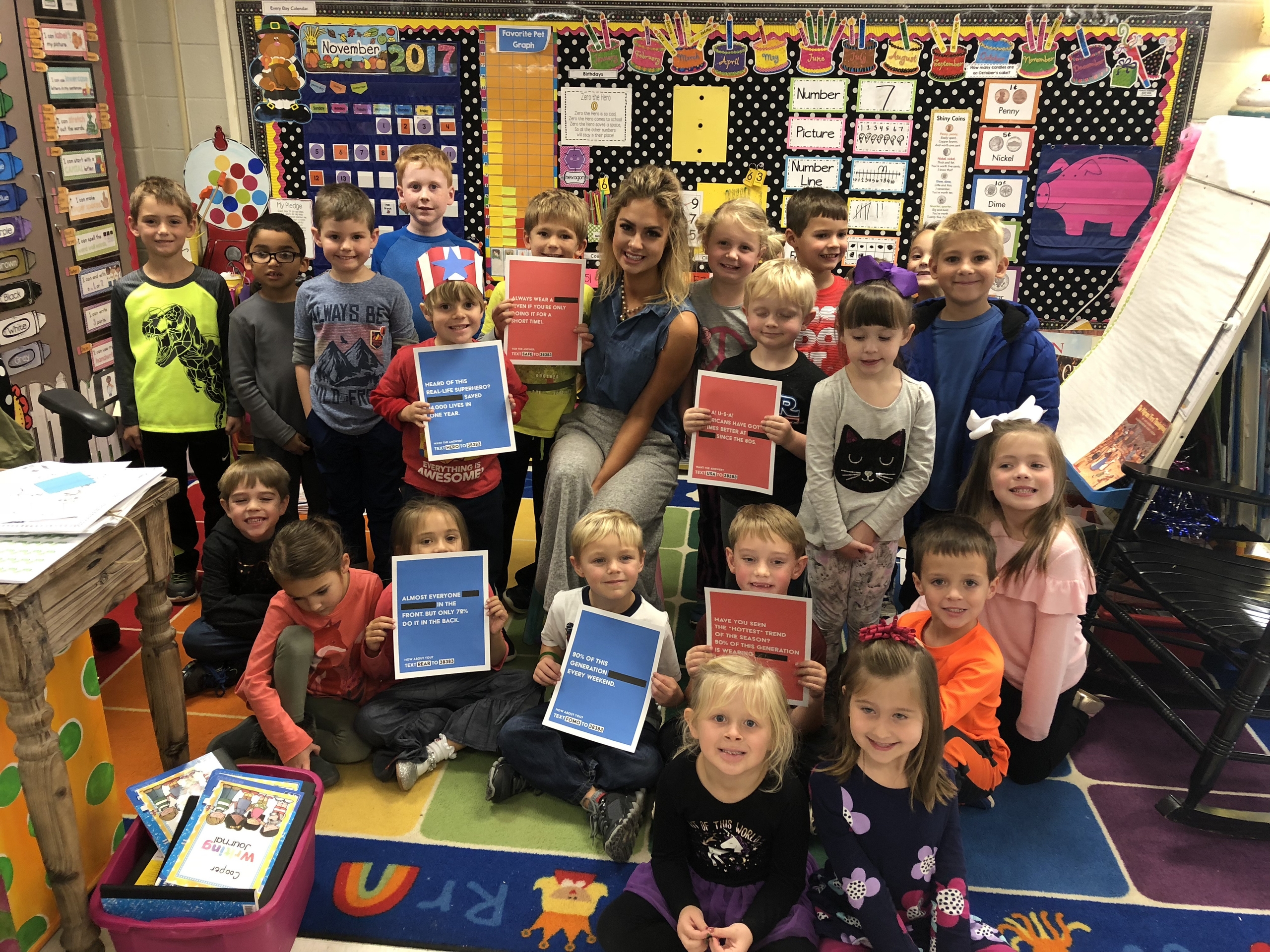 A diverse group of children and an adult educator gather in a classroom, smiling while holding colorful flyers promoting civic engagement and community service opportunities. The vibrant classroom is decorated with educational materials and artwork, emphasizing a supportive learning environment.