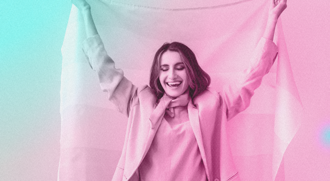 Young trans person, widely smiling, with long loose hair, wearing a blazer and blouse, with arms raised in joy