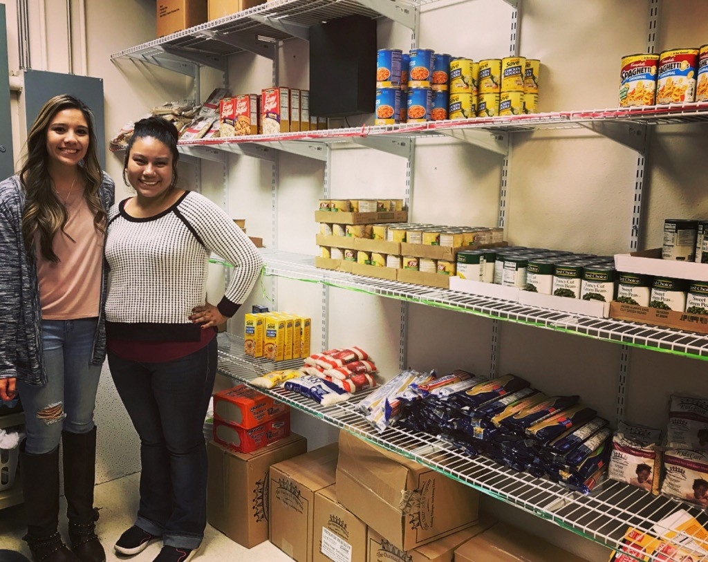 Two individuals stand in front of shelves stocked with various canned goods and dry food items in a community resource space. The image highlights volunteer engagement in providing essential food supplies for those in need.