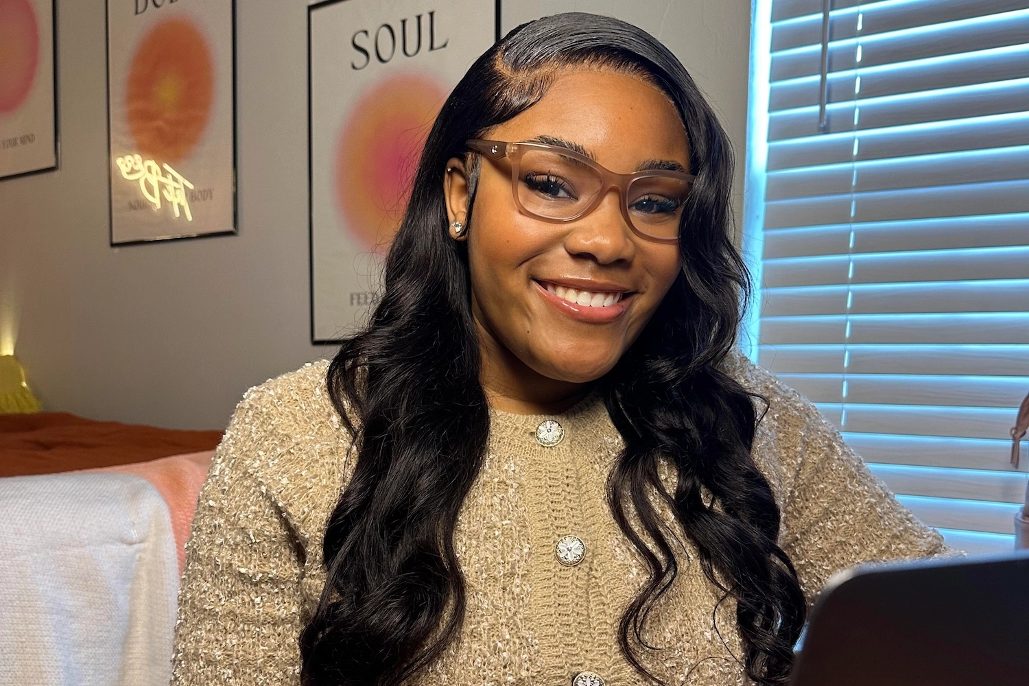 A smiling individual with styled dark hair and glasses is sitting at a desk, wearing a light-colored, textured sweater. Behind them are posters with colorful designs, highlighting a welcoming and creative environment for community service and civic leadership programming.