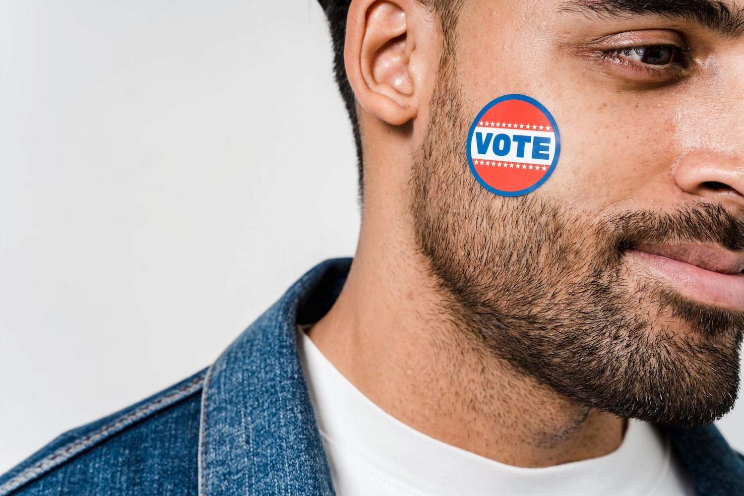 A young adult wearing a denim jacket, with a red, white and blue 'Vote' sticker on their face.