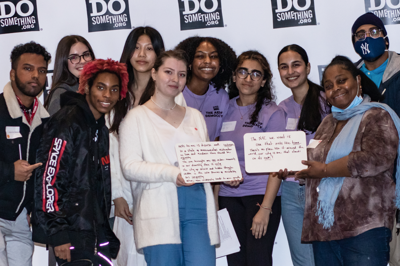A diverse group of individuals holds whiteboards with handwritten messages about community and belonging at a volunteer event hosted by DoSomething.org. The participants, representing various backgrounds, showcase their commitment to civic engagement and social change.