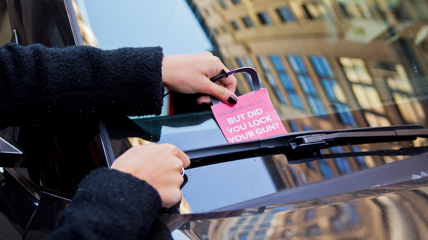 A person sticking a sign that reads But Did You Lock Your Gun under the windshield wiper of a car