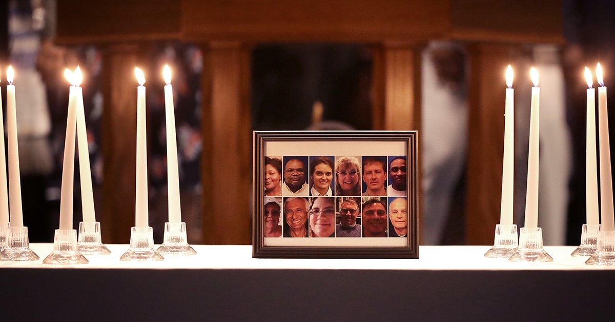 A framed collage of diverse faces displayed on a table surrounded by flickering white candles. The setting reflects a somber and respectful atmosphere, highlighting community connections and remembrance.