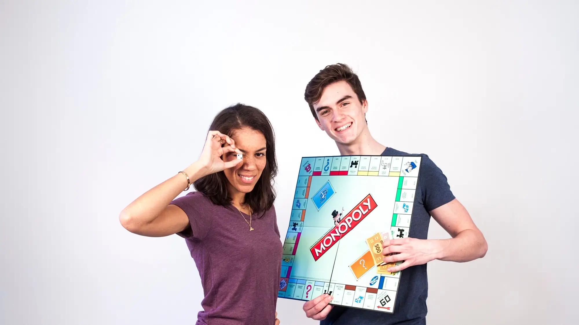 Two young people holding up for the camera a Monopoly board game and its pieces