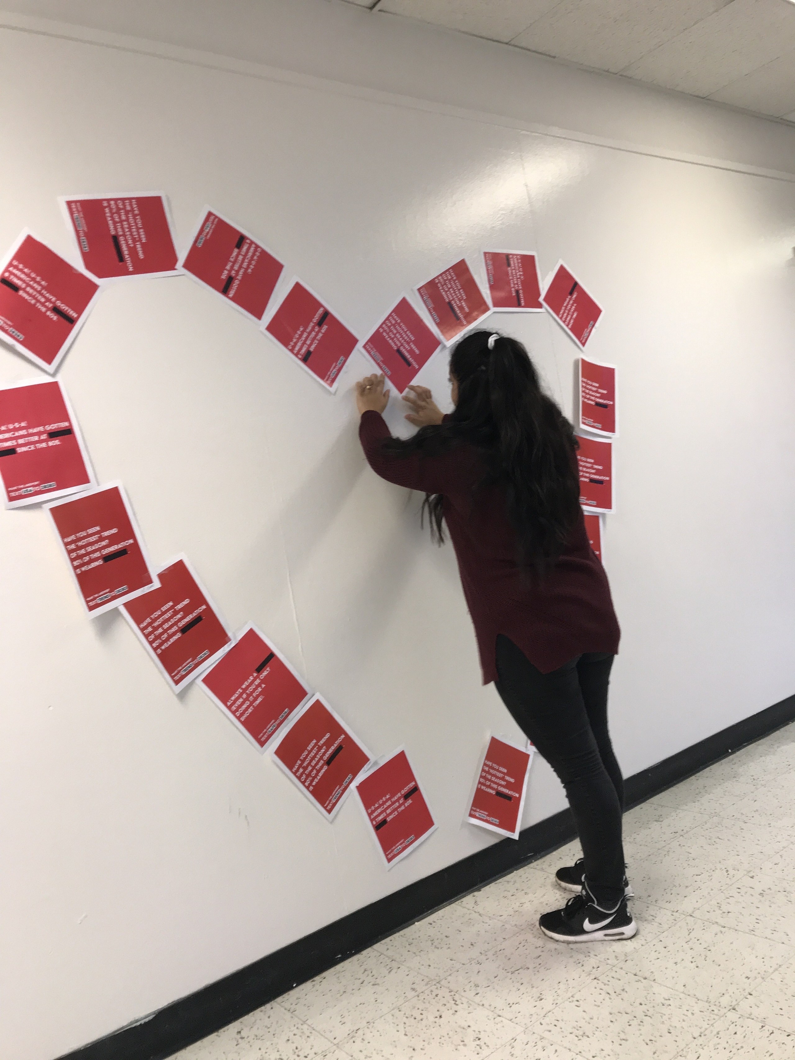 A person is attaching colorful flyers to a wall, creating a heart shape out of community service and civic engagement messages. The vibrant red flyers promote various volunteer opportunities and leadership programming.