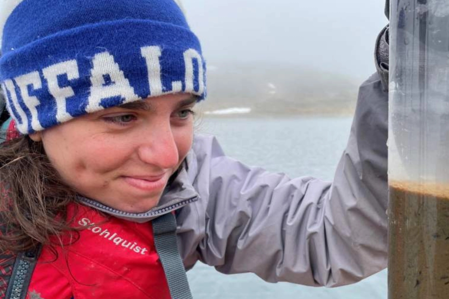 A person wearing a blue beanie and a red jacket closely examines a transparent cylinder filled with water and sediment, possibly during a community service or environmental research activity. The misty background suggests a natural outdoor setting, enhancing the focus on civic learning and volunteerism.