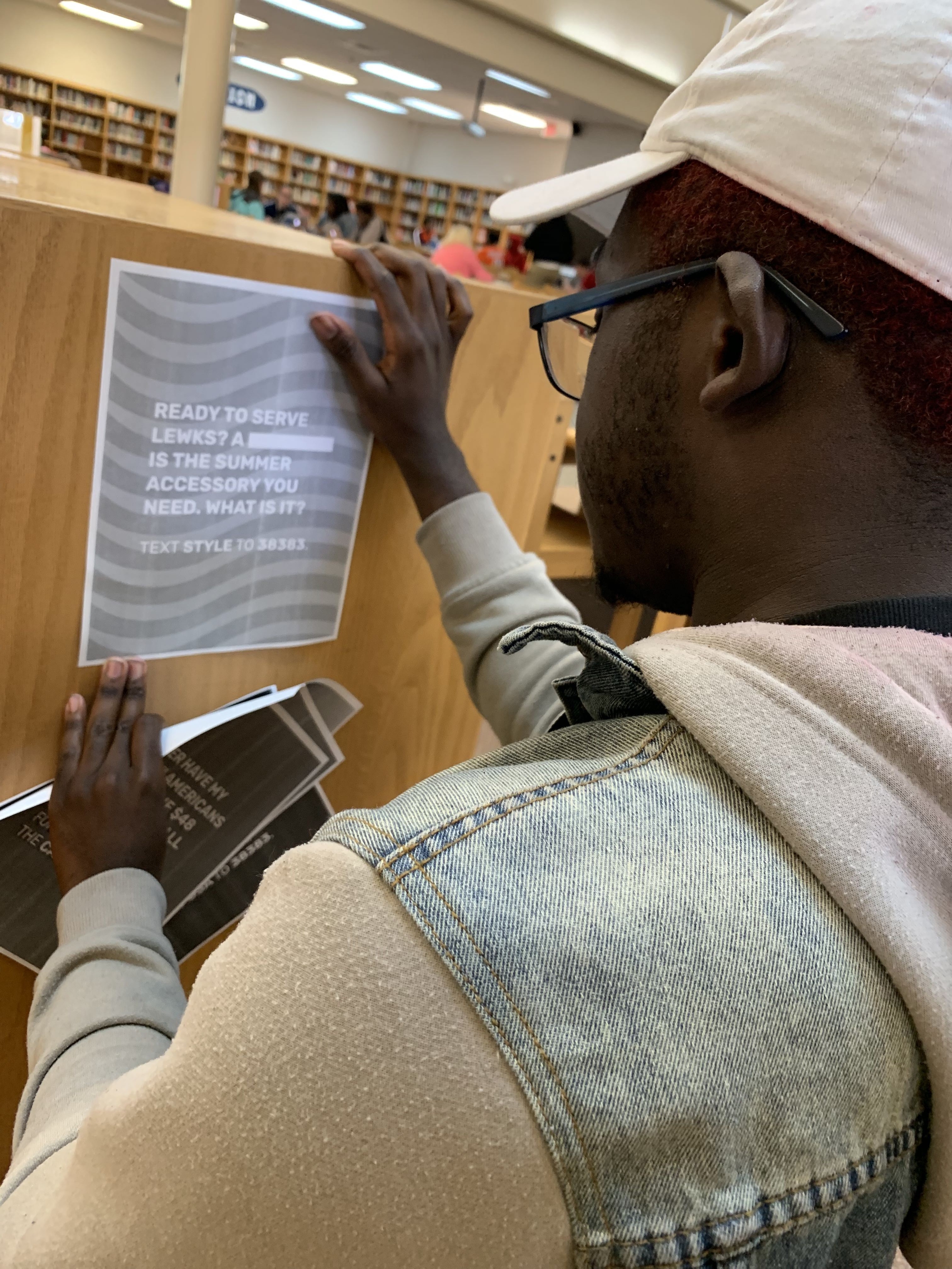 A person is putting up a flyer on a wooden divider in a library, promoting summer volunteer opportunities. The background shows community members engaged in various activities within the library setting.