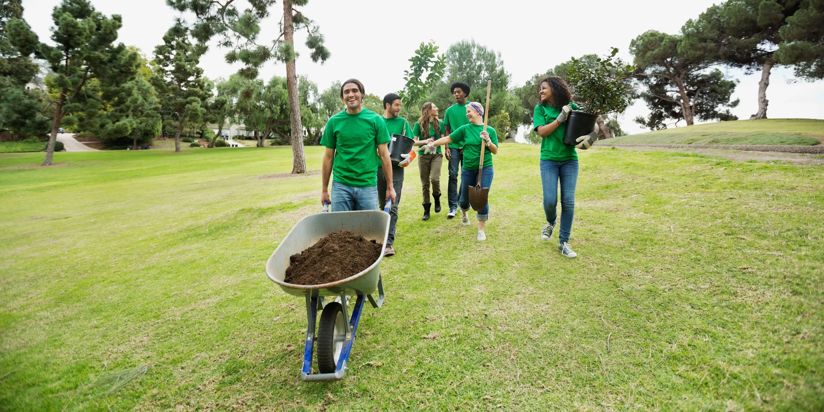 Six young adults volunteer to clean up a garden. One pushes a wheelbarrow, while others carry shovels and gardening supplies needed for the community cleanup.