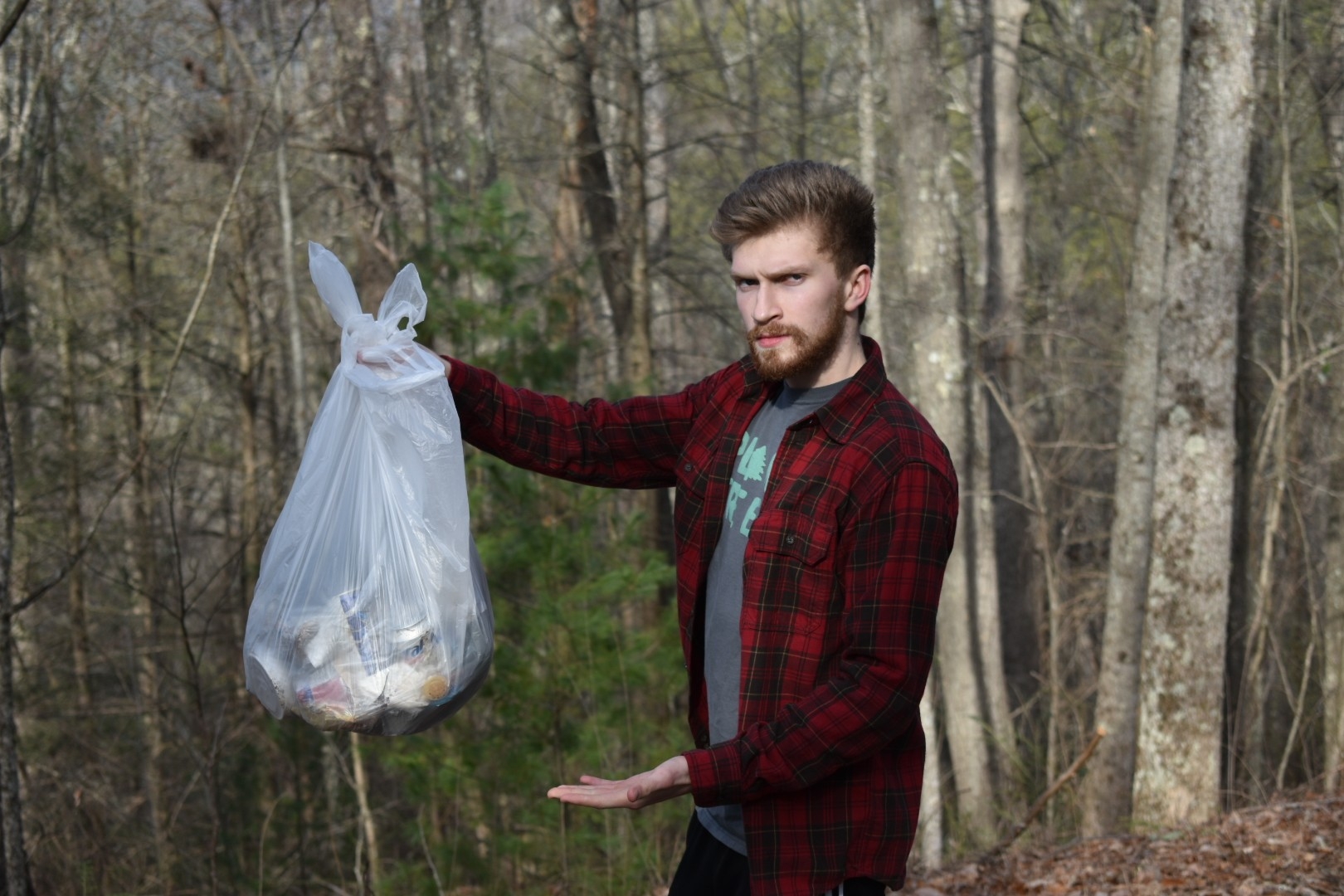 A person standing in a forest holds up a plastic bag filled with trash, promoting environmental awareness and community service. The surrounding trees emphasize the importance of maintaining a clean natural environment.