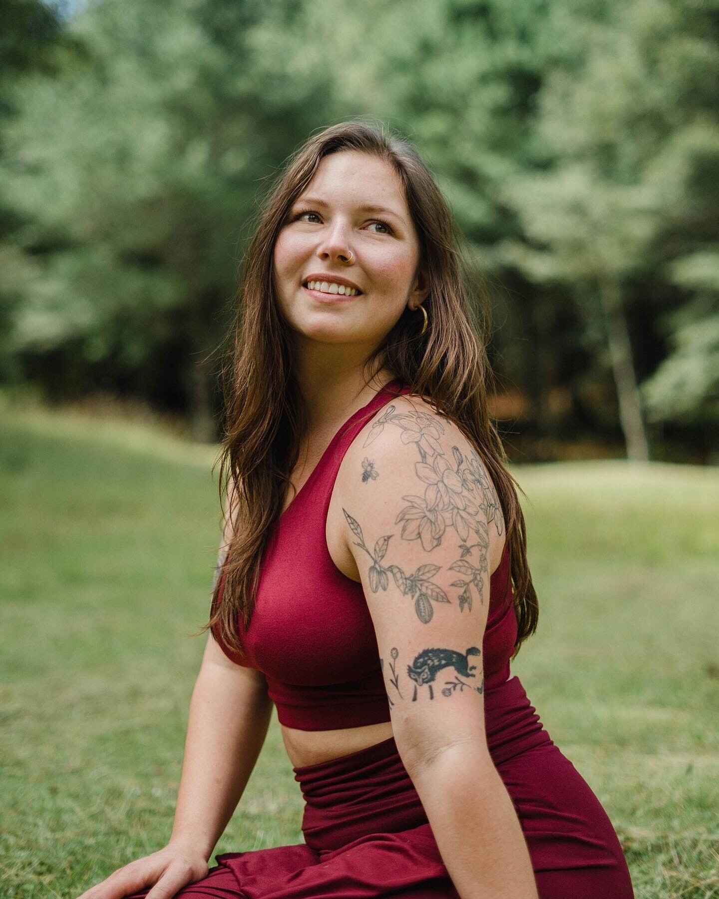 A person with long hair sitting in a green outdoor setting, wearing a red crop top and skirt, smiling while looking thoughtfully to the side. Visible tattoos decorate their arm, featuring floral and animal designs, illustrating a connection to nature.