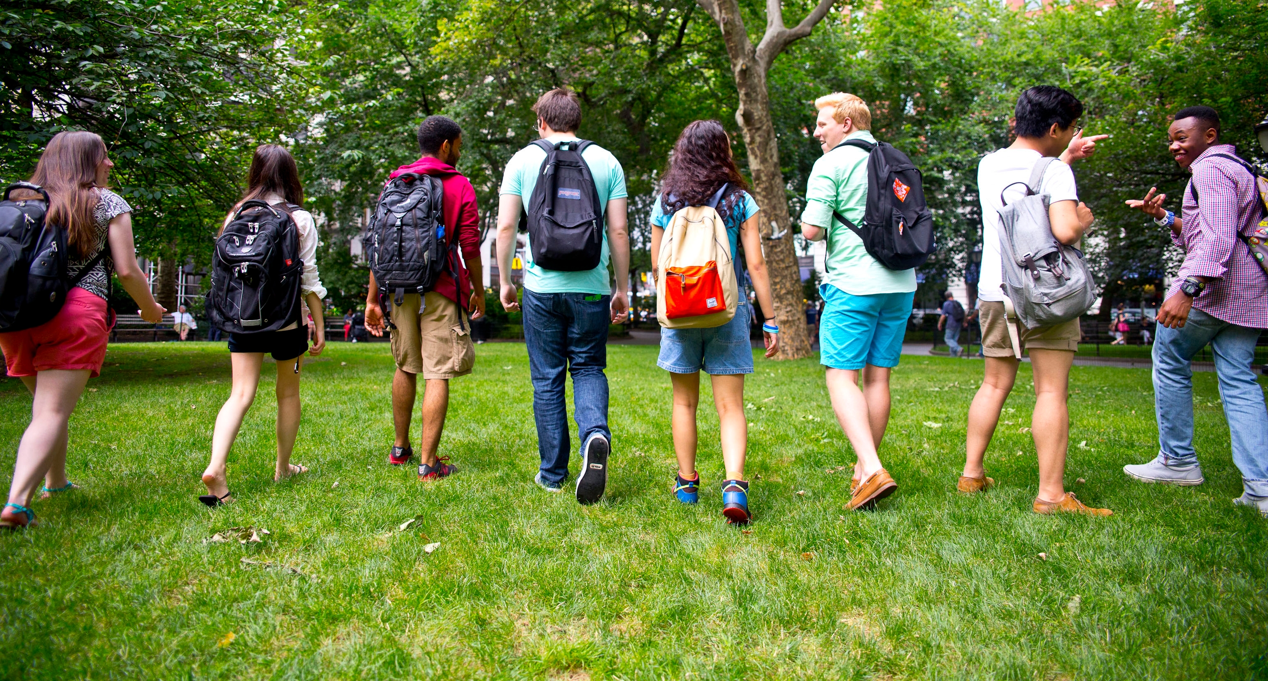 A group of eight people walking across a grassy tree-lined area with their backs to the camera, talking together, all wearing backpacks of various styles and colors