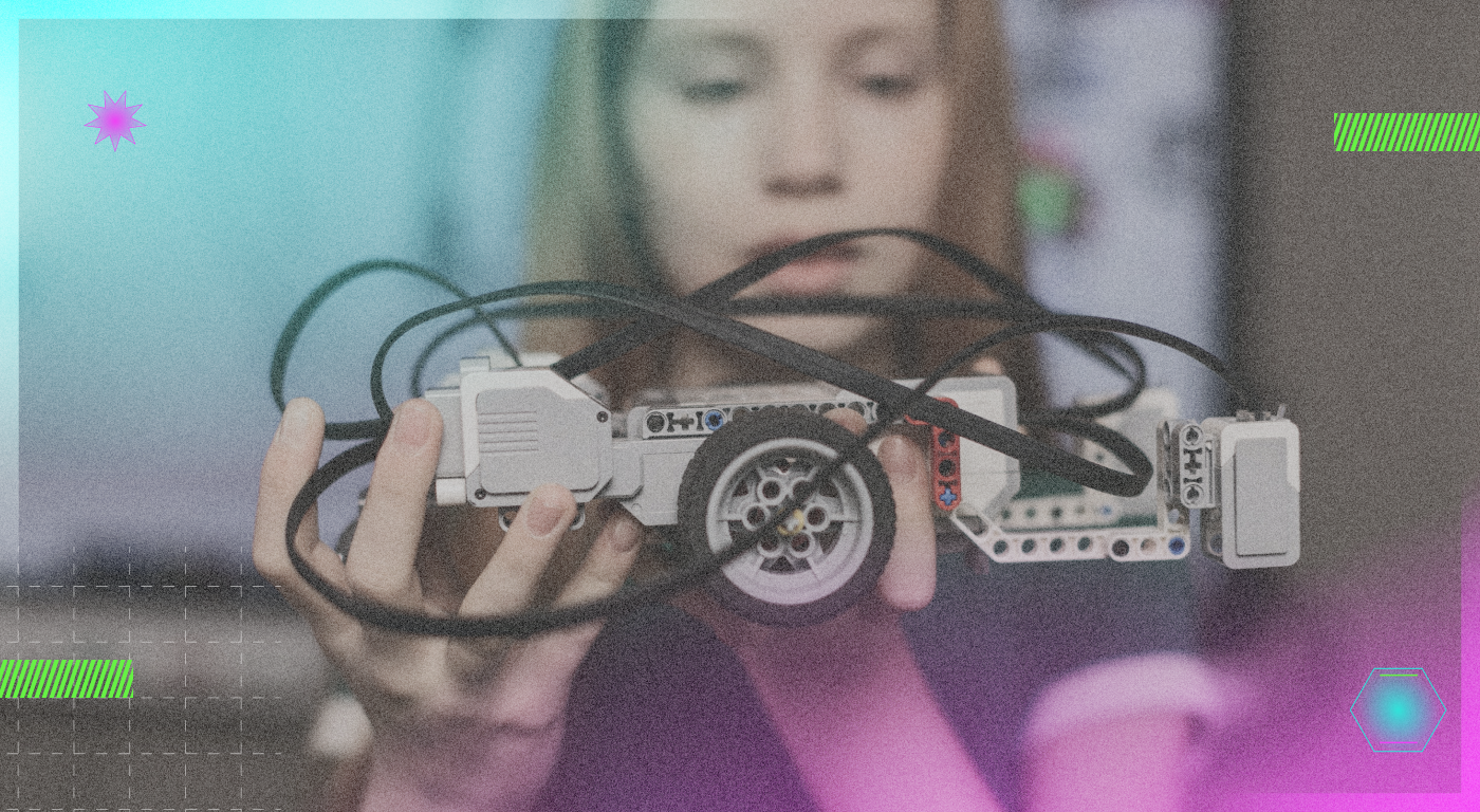 A young person with long light colored hair holding a small wheeled robot with several loose cables