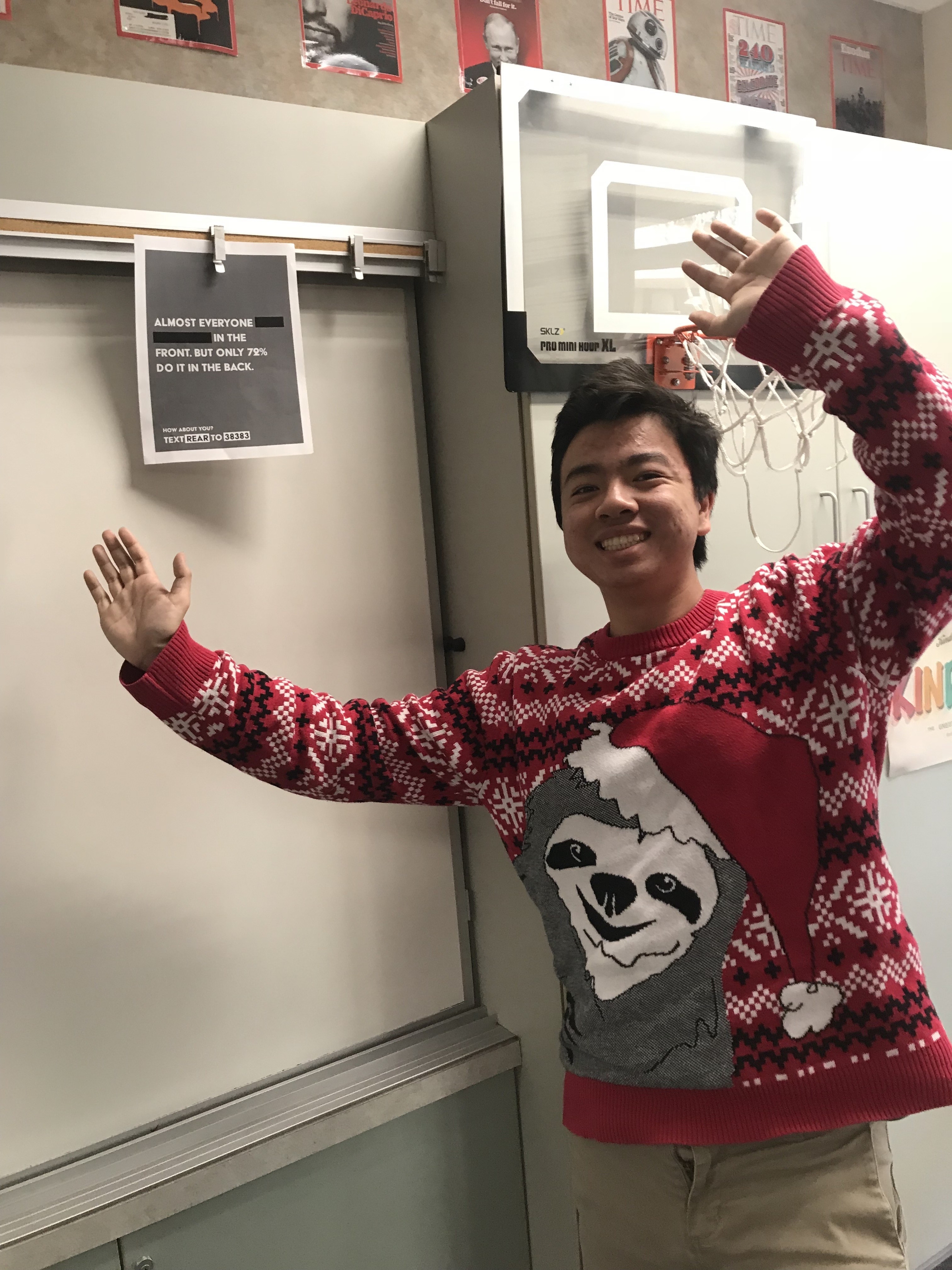 A cheerful individual wearing a festive red and white Christmas sweater featuring a sloth and a Santa hat, stands beside a basketball hoop in a classroom. A poster on the wall reads about a statistic related to participation, enhancing the atmosphere of community and engagement.