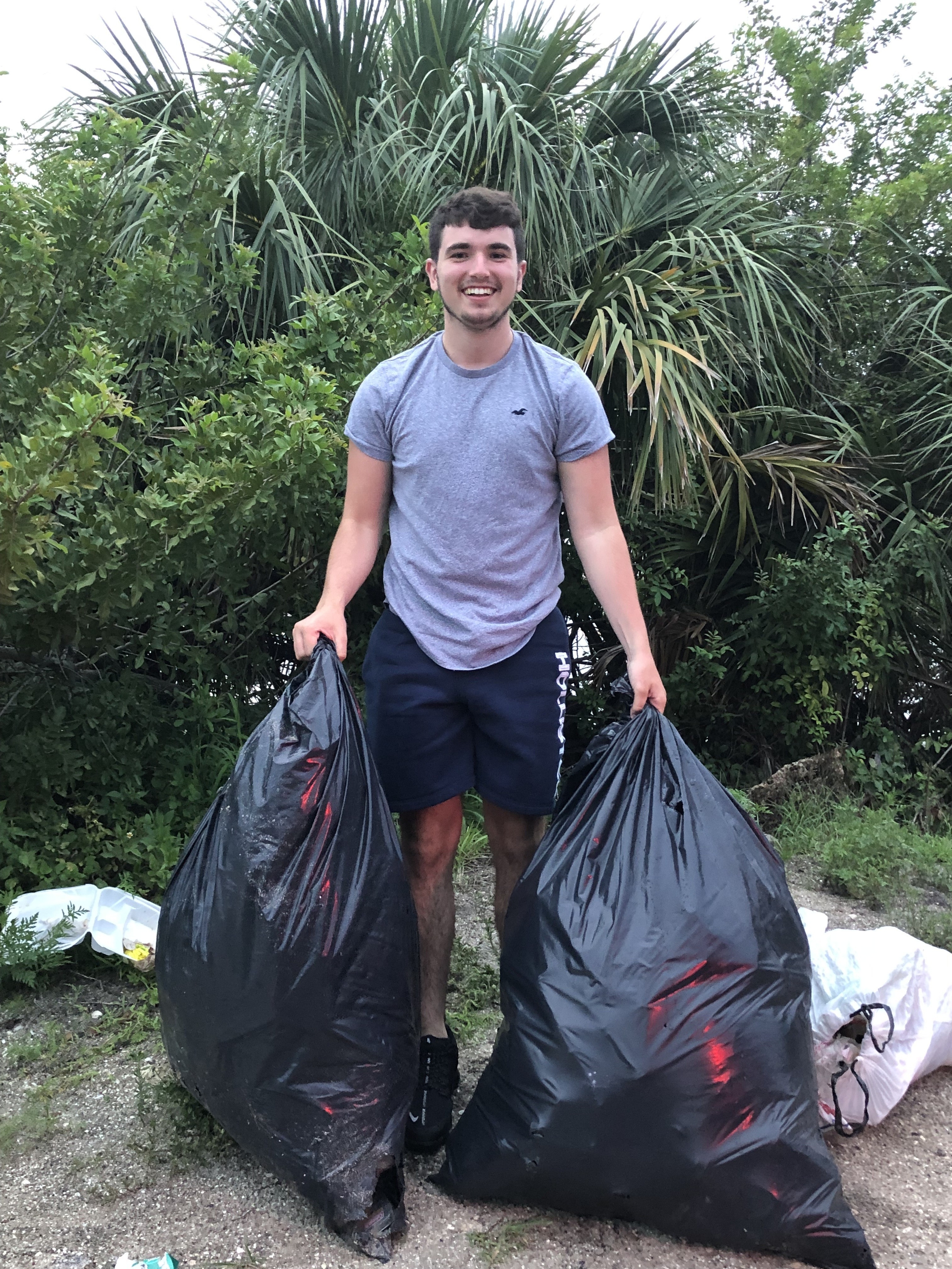 An individual stands outdoors, smiling and holding two large black trash bags filled with litter, surrounded by lush greenery. This image highlights community service efforts focused on environmental cleanup and volunteer engagement.