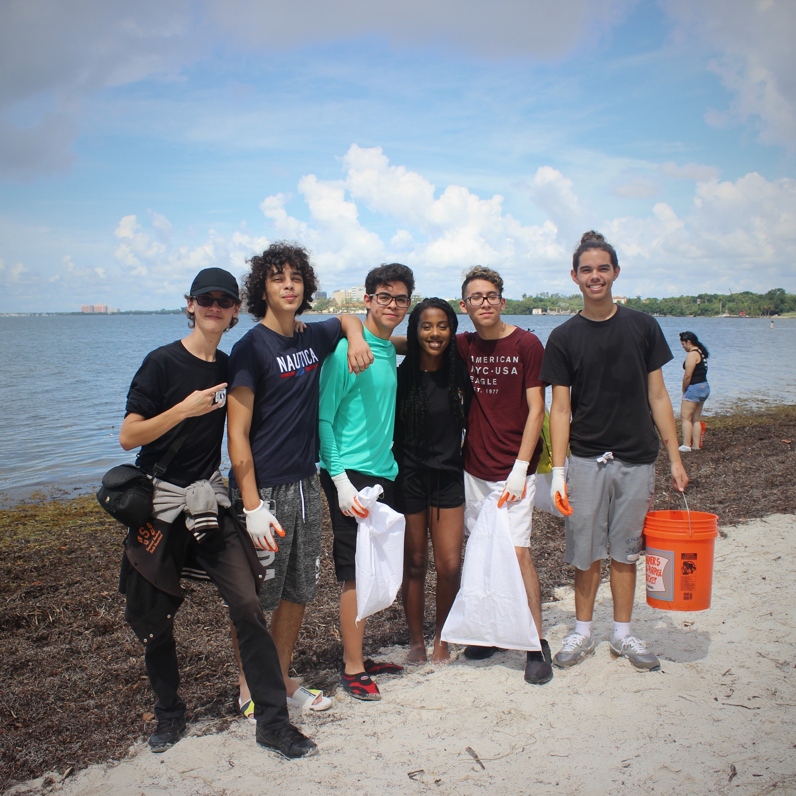 A diverse group of six individuals stands on the beach holding trash bags, smiling while participating in a community cleanup project by the water. The scene highlights volunteerism and civic engagement as they contribute to environmental sustainability.