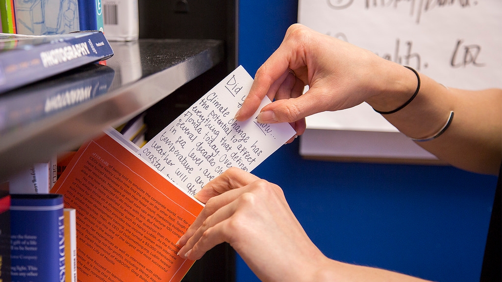 A person slipping a home made bookmark with facts about climate change into a book that rests on a bookshelf