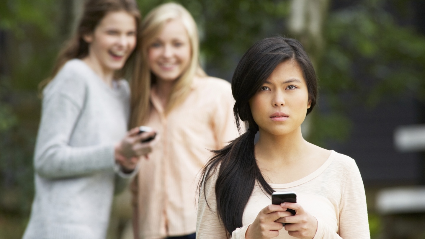 Young woman with a concerned expression on her face, holding her cellphone, with two women in the background slightly blurred laughing