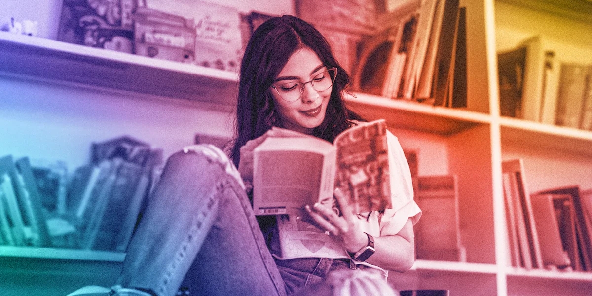 Cool to warm tone gradient of young person with long dark hair and cateye wireframe glasses reading a book with book shelves in the background