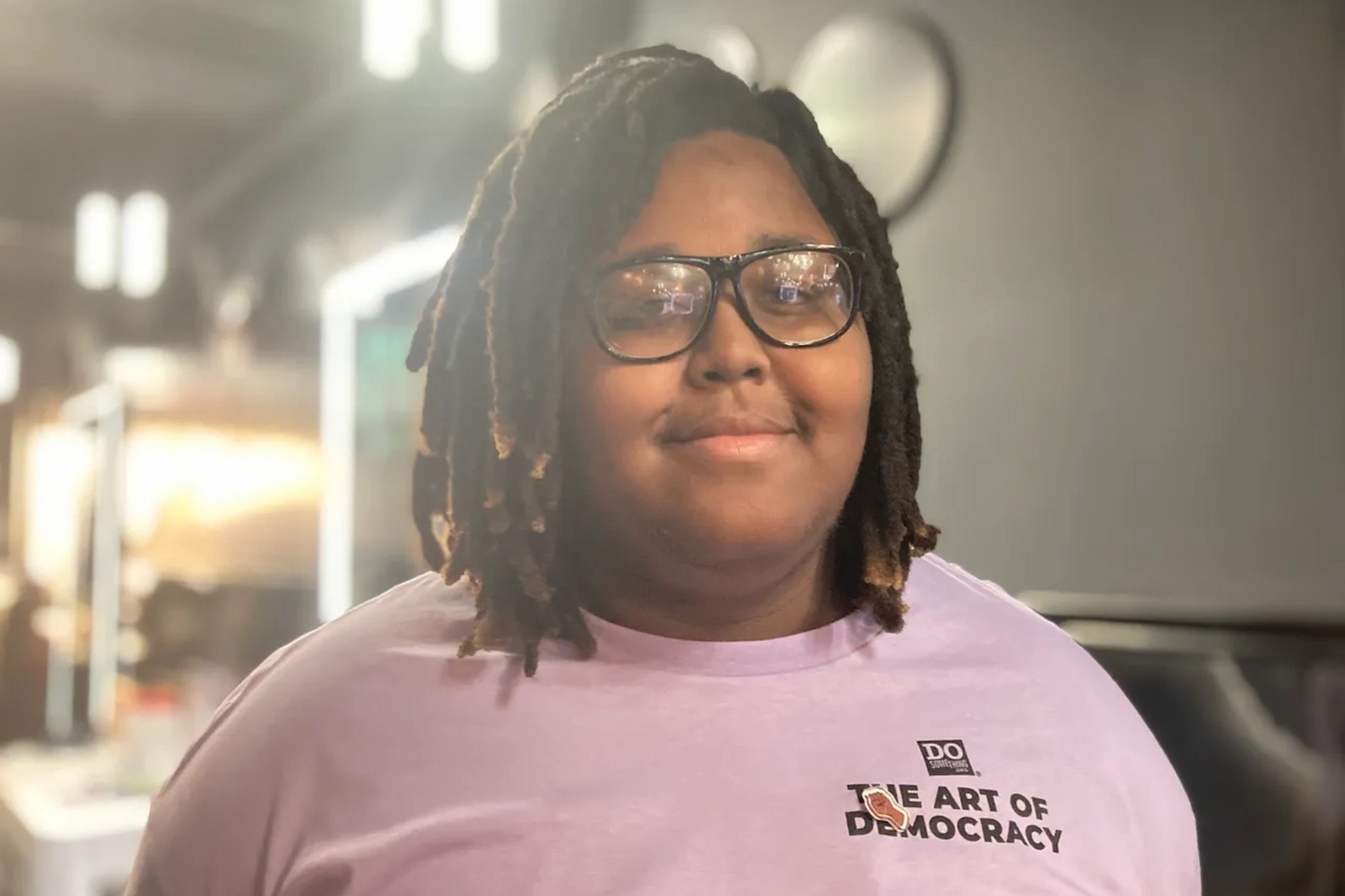 A young person with black glasses, wearing a lavender DoSomething Art of Democracy t-shirt, and pins.