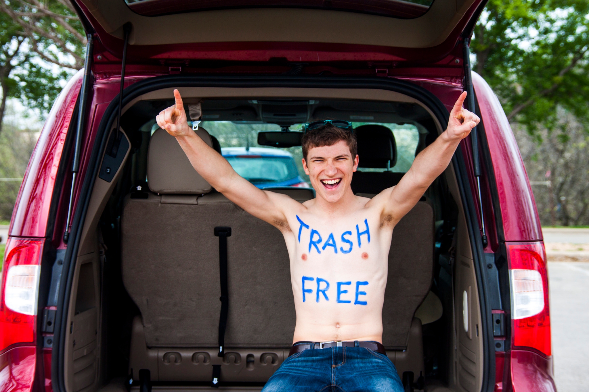 Young person shirtless with arms raised, sitting on a minivan's tailgate, with "trash free" painted on their chest