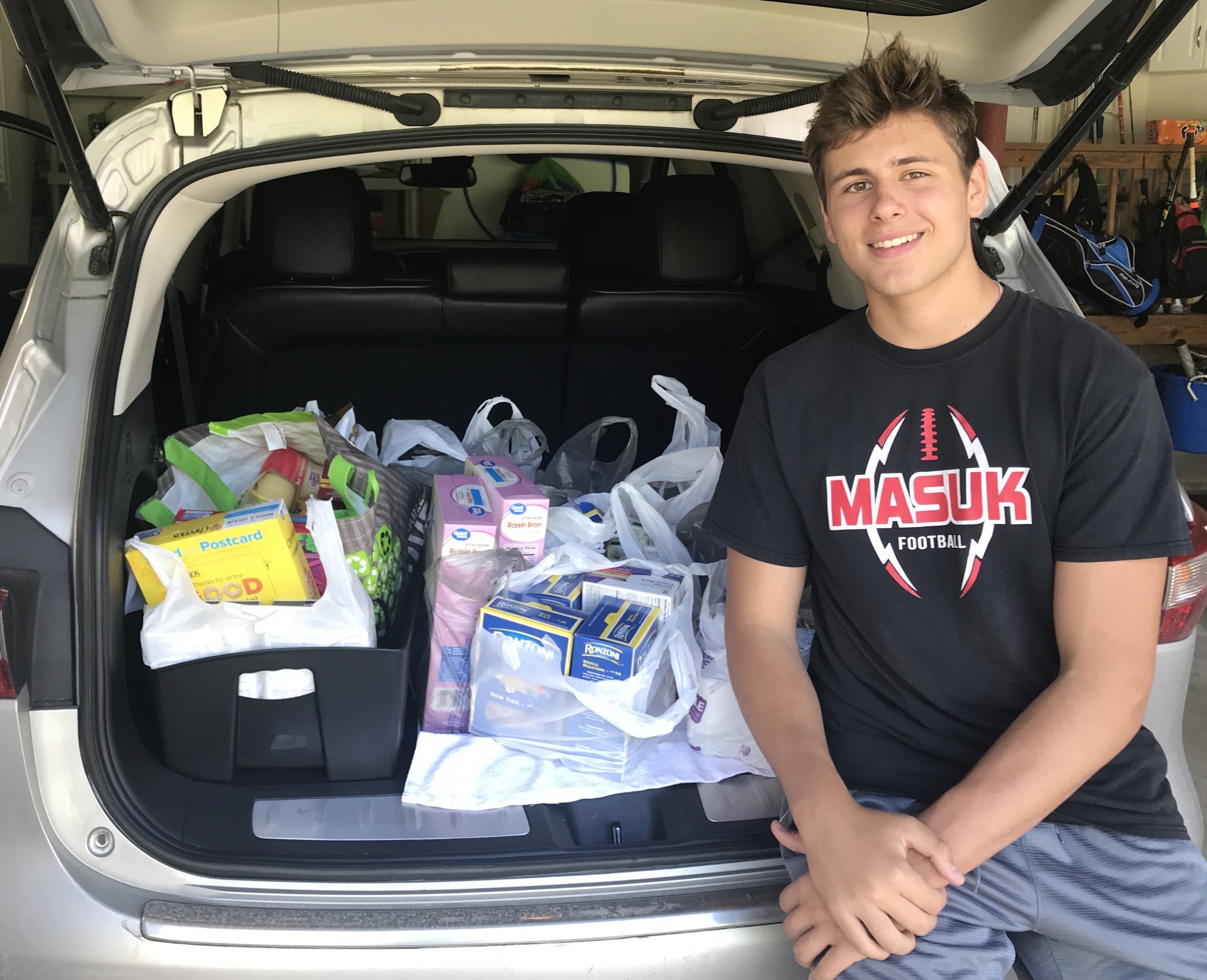 A young volunteer sits beside a car trunk filled with bags of groceries and supplies, showcasing community service efforts. This image highlights opportunities for civic engagement and the importance of giving back to the community.