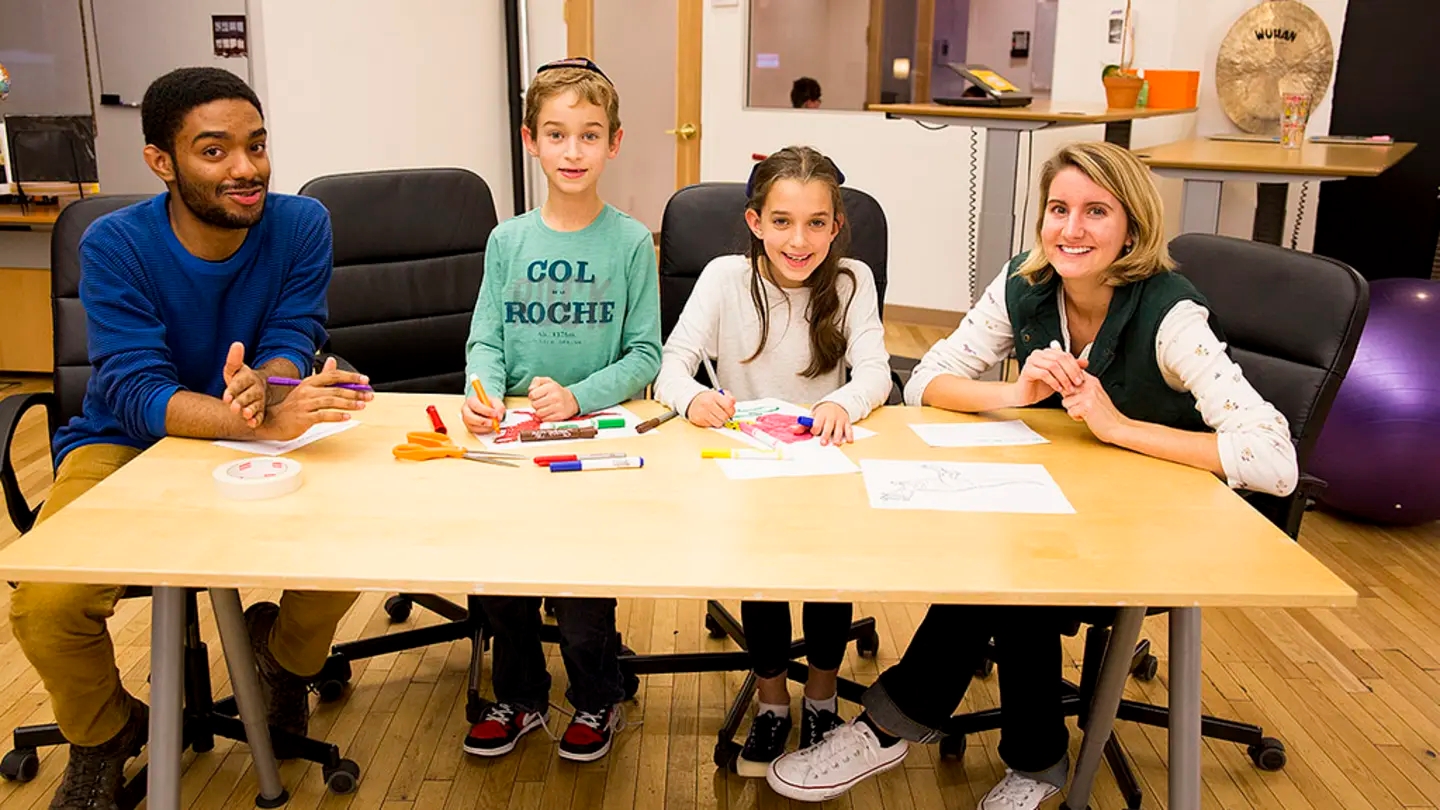 Two young people at a table with two children, with books and markers