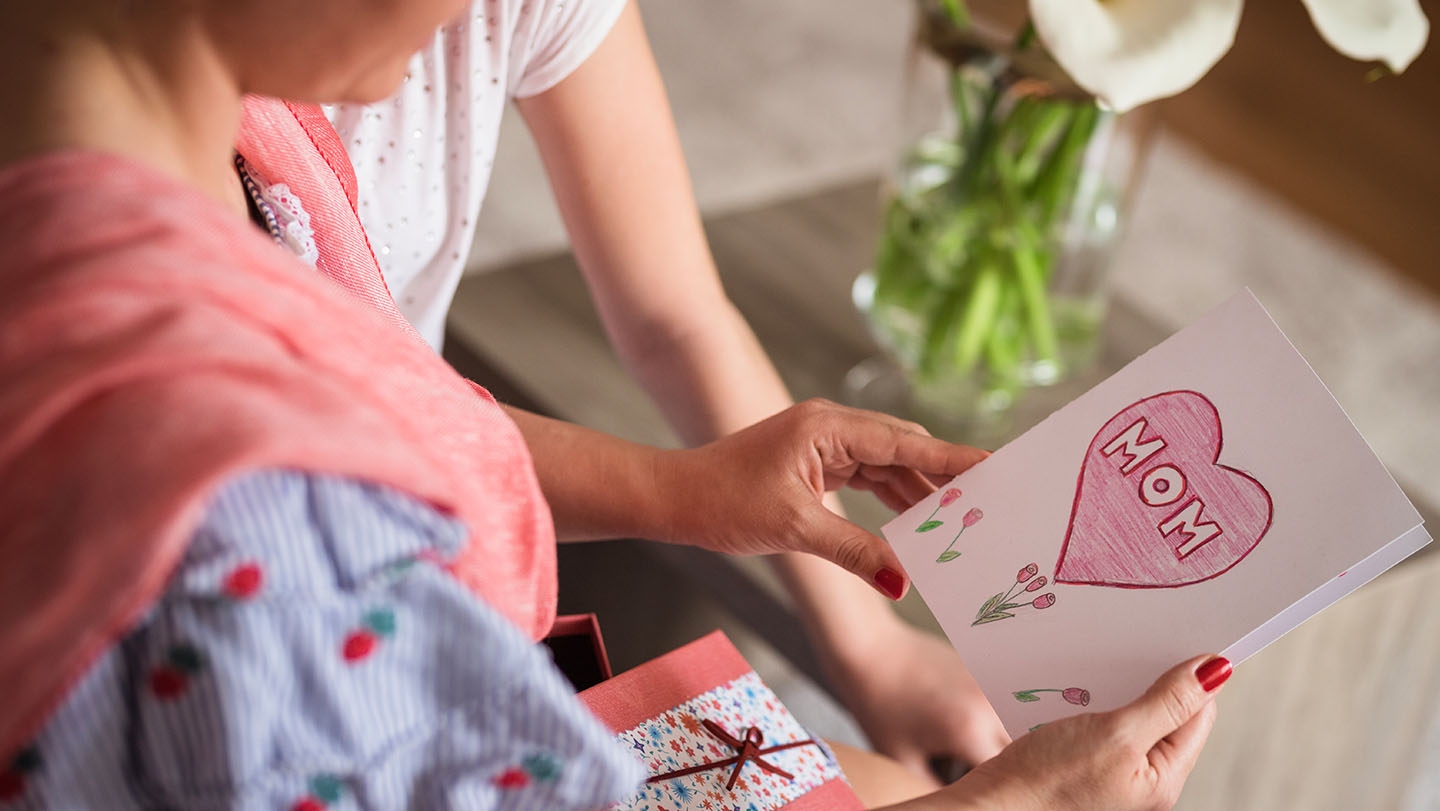 A close up of a mother holding a card that says "MOM" inside a handdrawn heart, a young child stands close to the mother