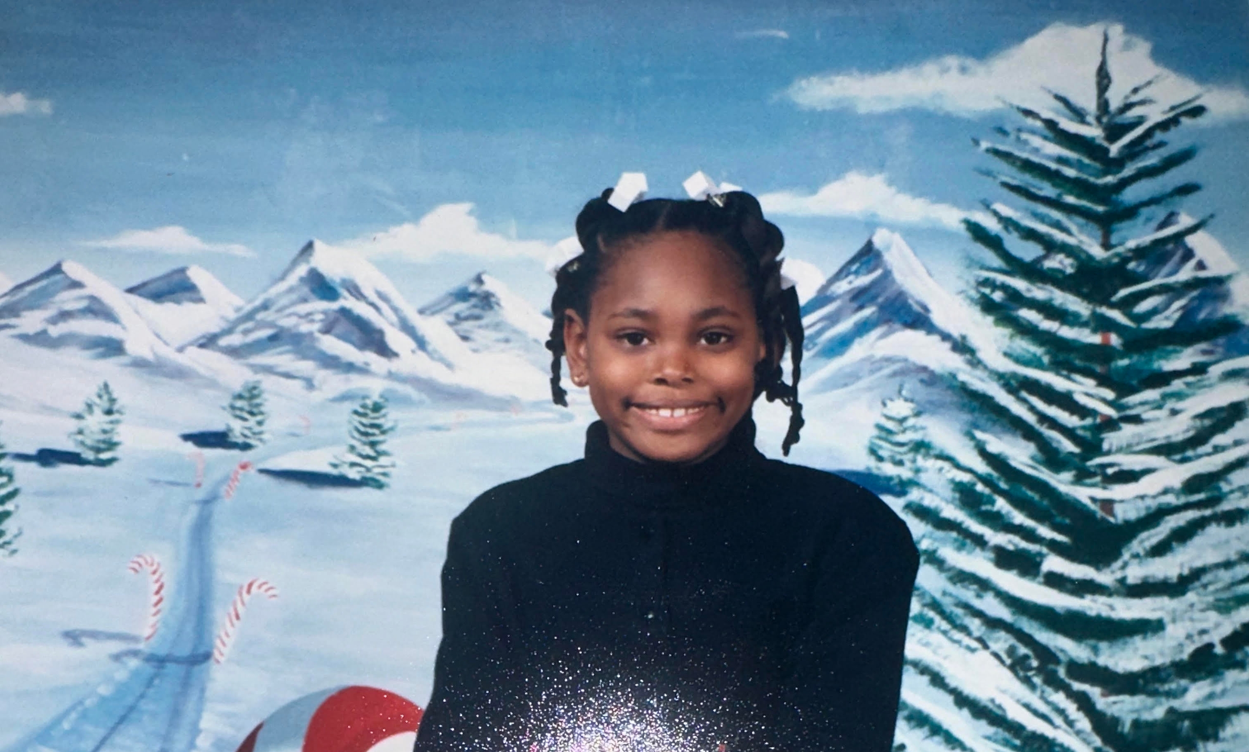 A smiling child with braided hair sits confidently on a red ladder, wearing a black long-sleeve shirt, a plaid skirt, and black boots, against a festive winter-themed backdrop featuring mountains, a snowy landscape, and holiday gifts. This image represents the joy of community engagement and the spirit of volunteerism.