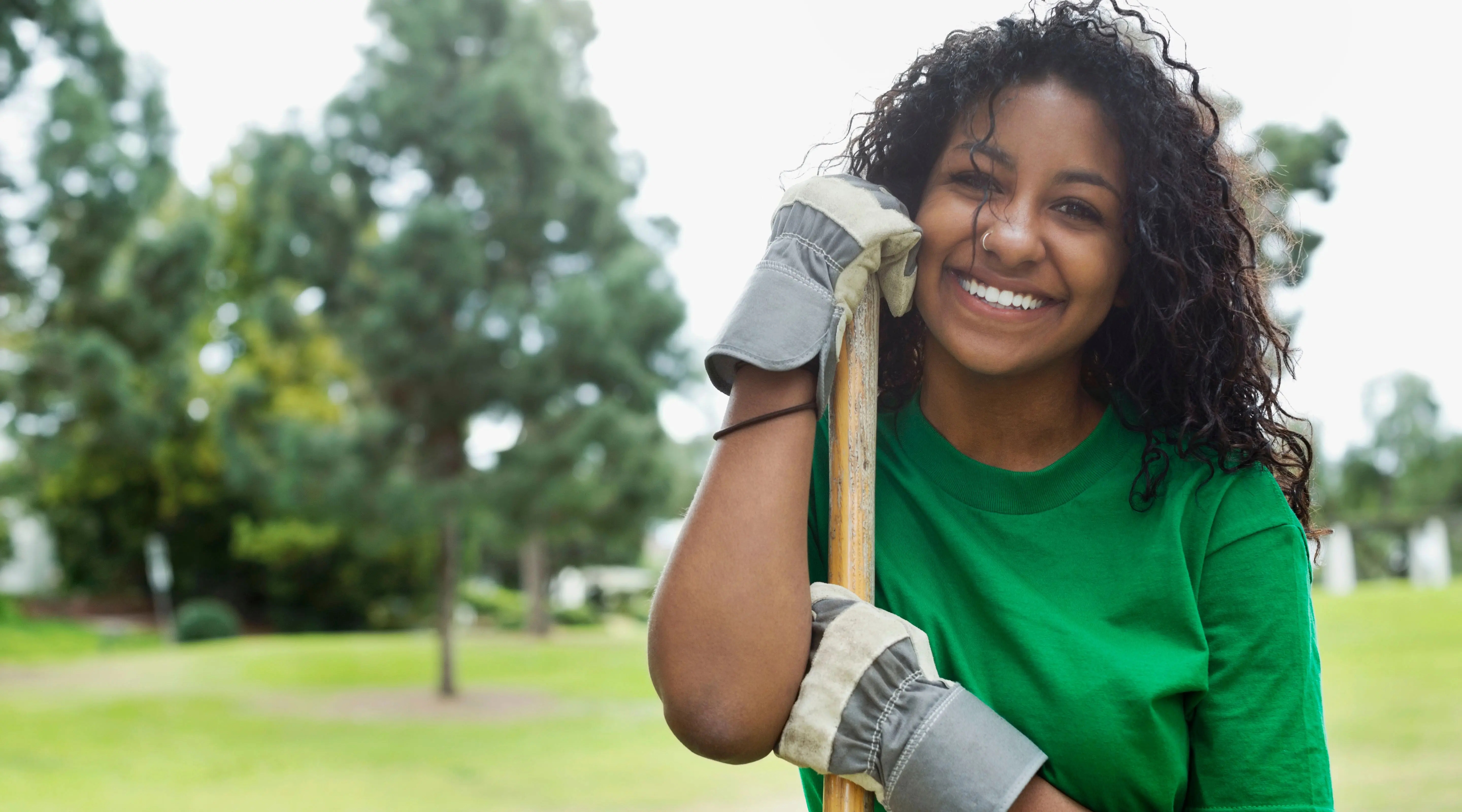 A young person poses in a park with a garden tool and gloves, signifying that they are working to make a community space more green.