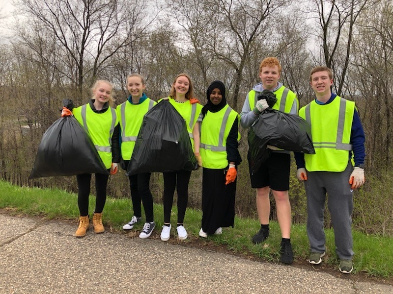 A diverse group of six volunteers in bright safety vests stand together on a grassy roadside, each holding large trash bags filled with collected litter. This image highlights community service and civic engagement efforts focused on environmental cleanup.