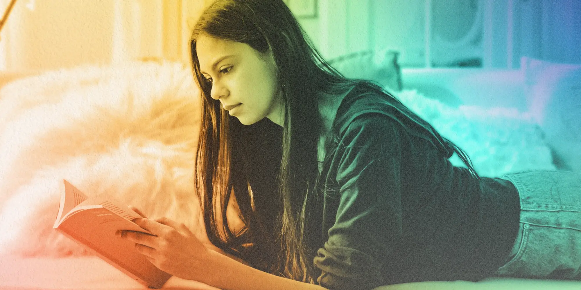 Warm to cool gradient of a young person with long hair and a zip up hoodie laying on their stomach and reading a book