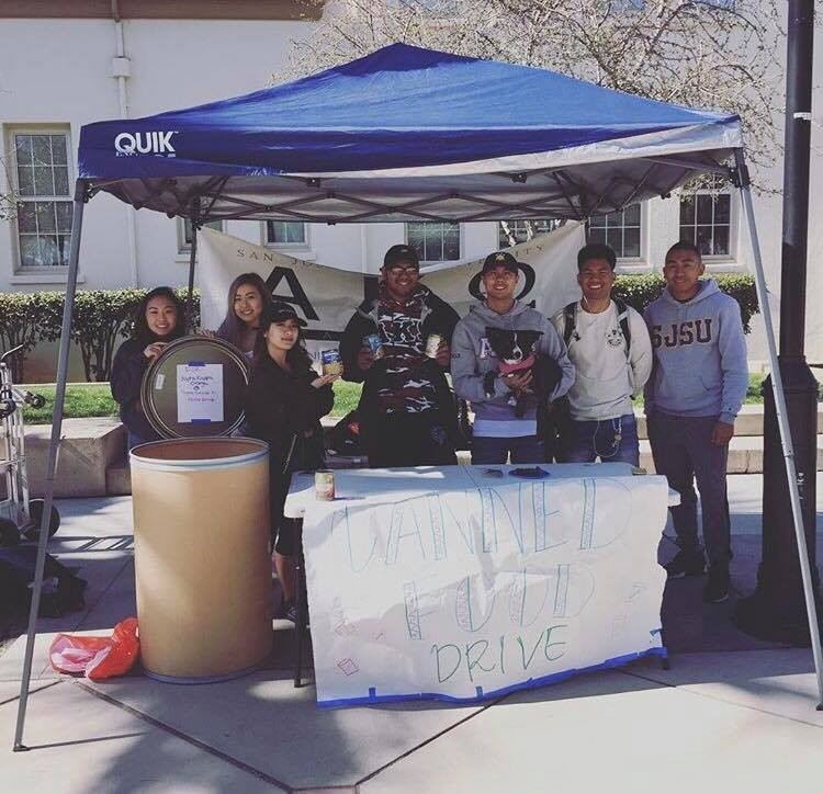 A diverse group of six volunteers stands behind a table at a food drive event, displaying cans and a sign that reads 'Canned Food Drive'. They are promoting community service and civic engagement under a blue canopy on a sunny day.