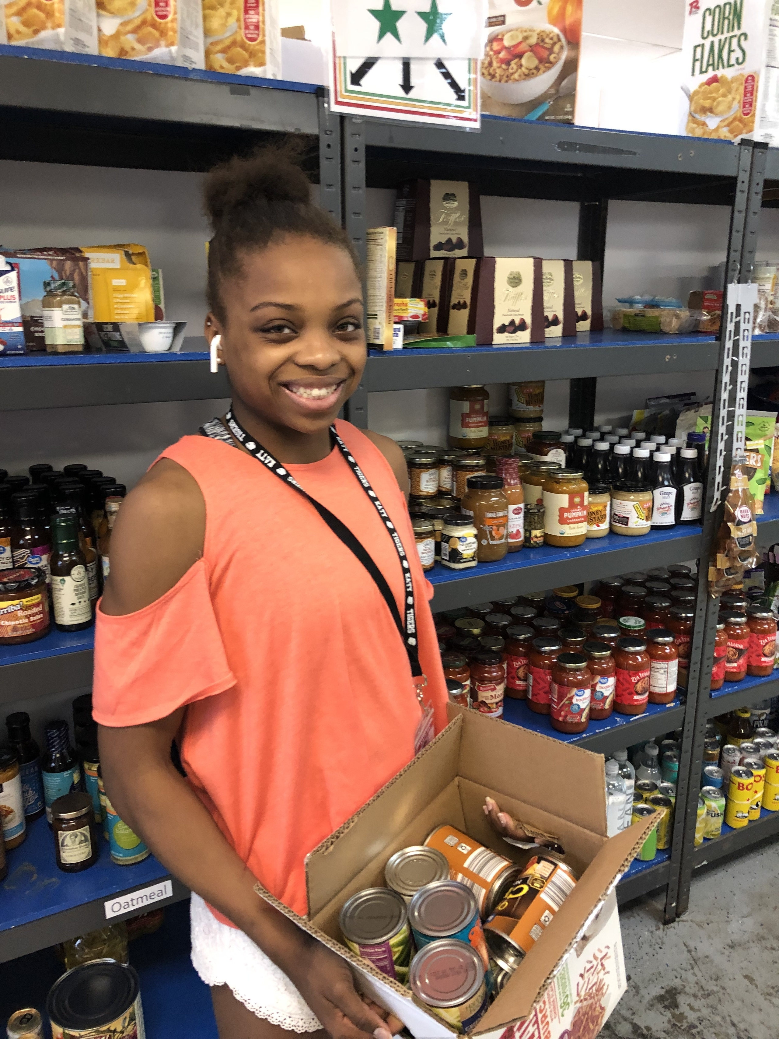 A volunteer in an orange shirt smiles while holding a box of canned goods in a community food pantry. Behind, shelves are filled with various food items, highlighting opportunities for community service and civic engagement.
