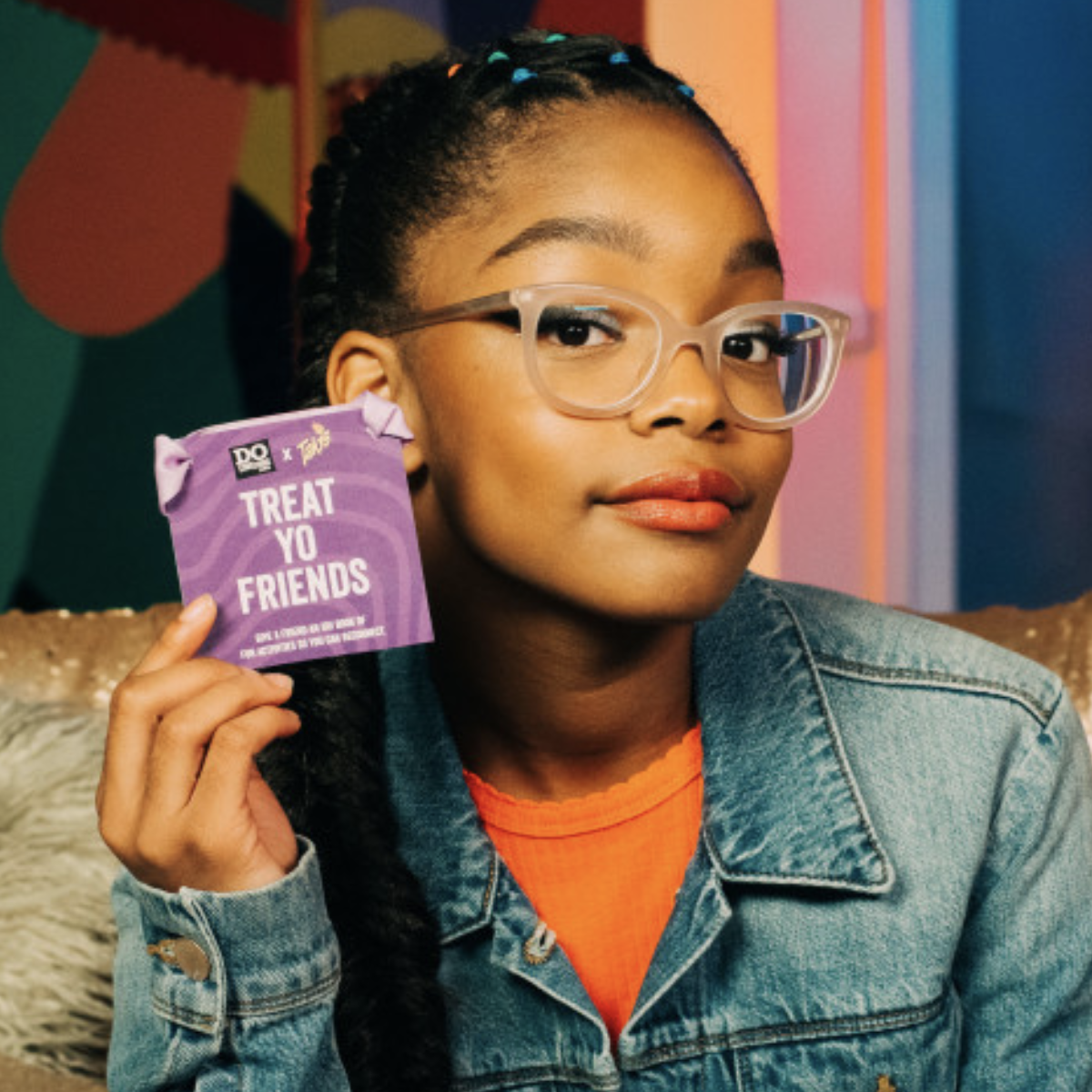 A young person with glasses poses confidently while holding a purple card that reads 'TREAT YO FRIENDS.' The image emphasizes community engagement and the spirit of sharing in volunteer and civic programs.