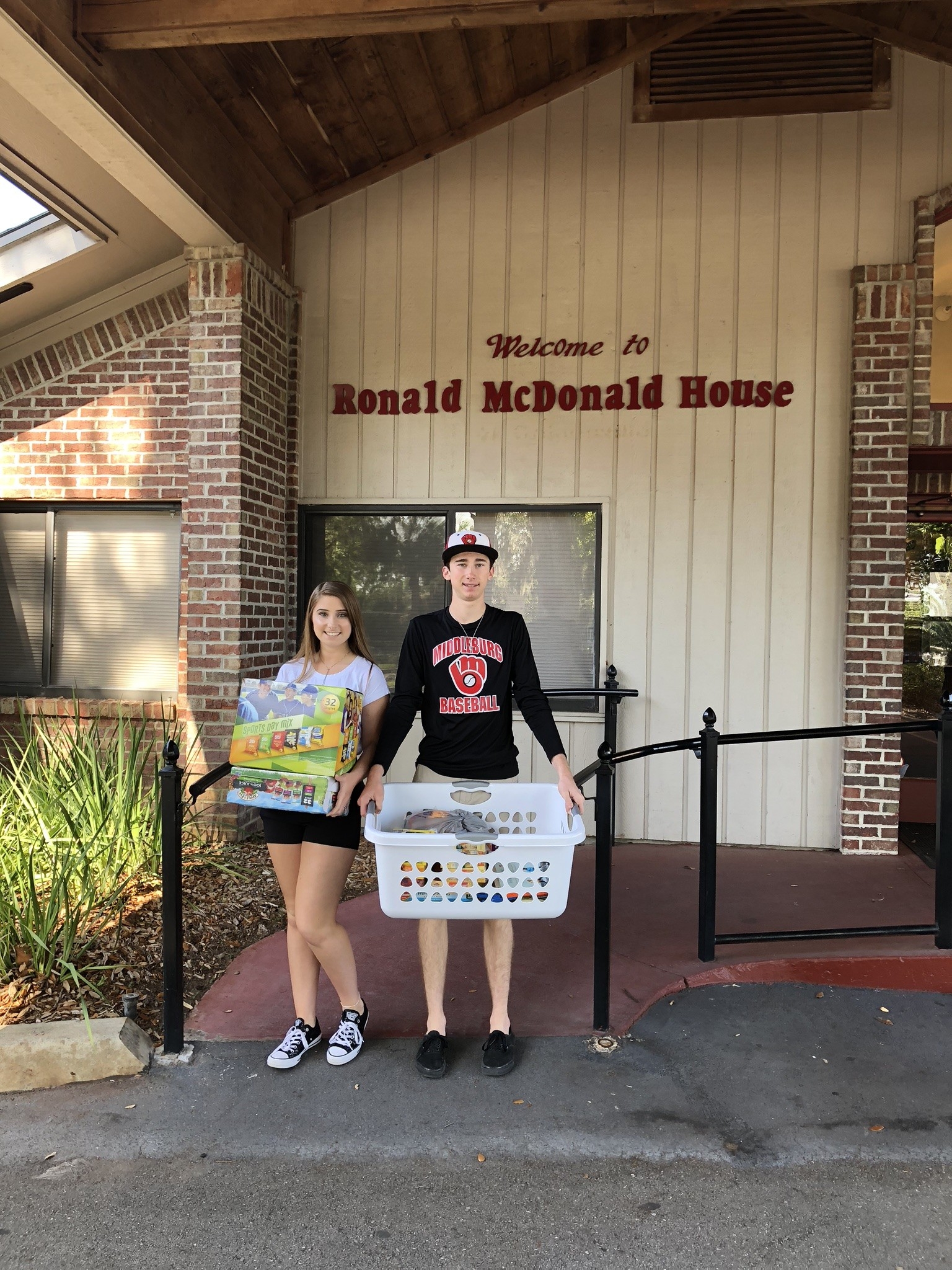 Two volunteers stand outside the Ronald McDonald House, one holding a box of supplies and the other carrying a laundry basket. This image highlights community service efforts and volunteer opportunities benefiting families in need.